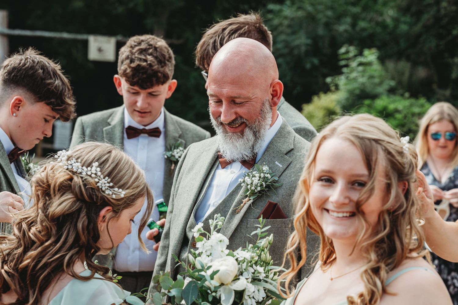 A joyful outdoor wedding scene with guests gathered around bridal bouquets featuring white flowers and greenery.