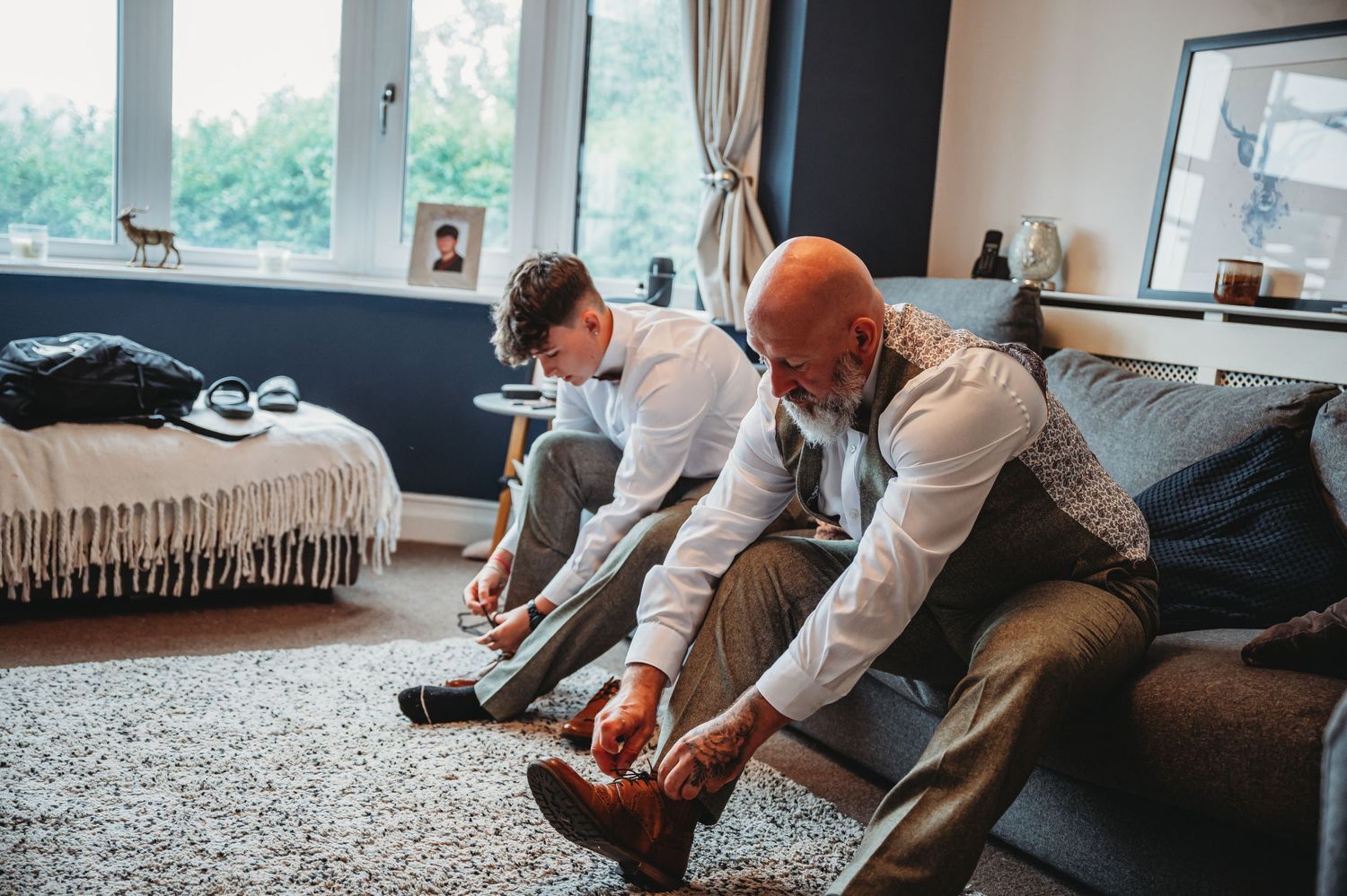 Two people in formal attire sit on a couch tying their dress shoes while getting ready for an event.