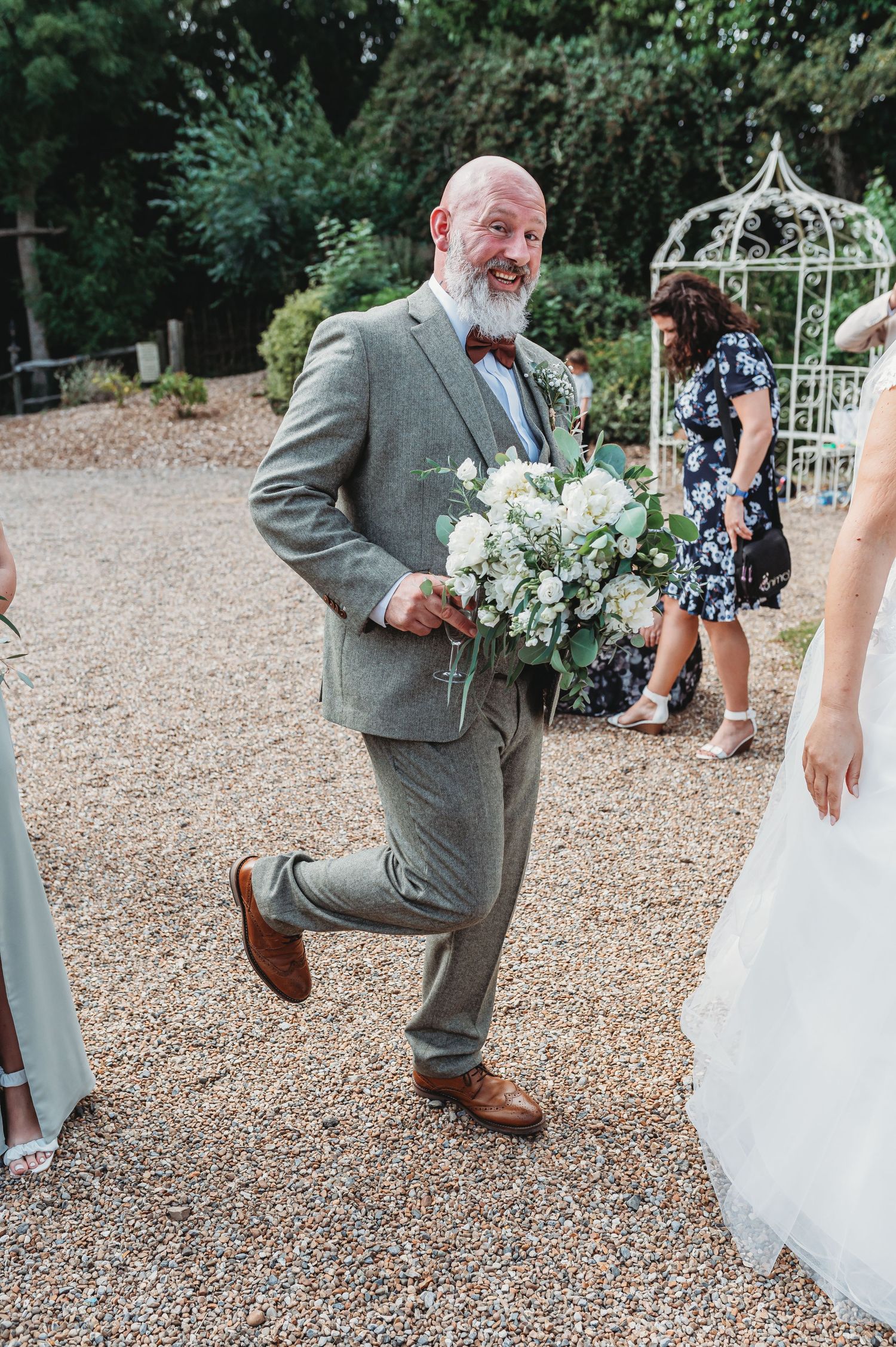 A man in a gray suit and bowtie dances playfully on a gravel path while holding a white floral bouquet at an outdoor wedding.