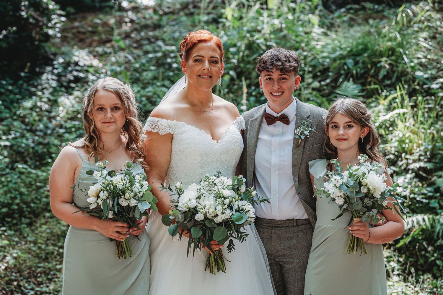 Wedding party poses together outdoors in sage green and white attire holding floral bouquets in a garden setting.