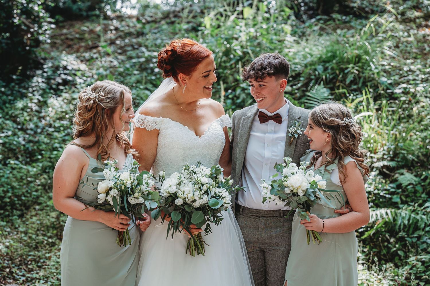 A bridal party holds lush greenery and white flower bouquets while posing together in a garden setting.