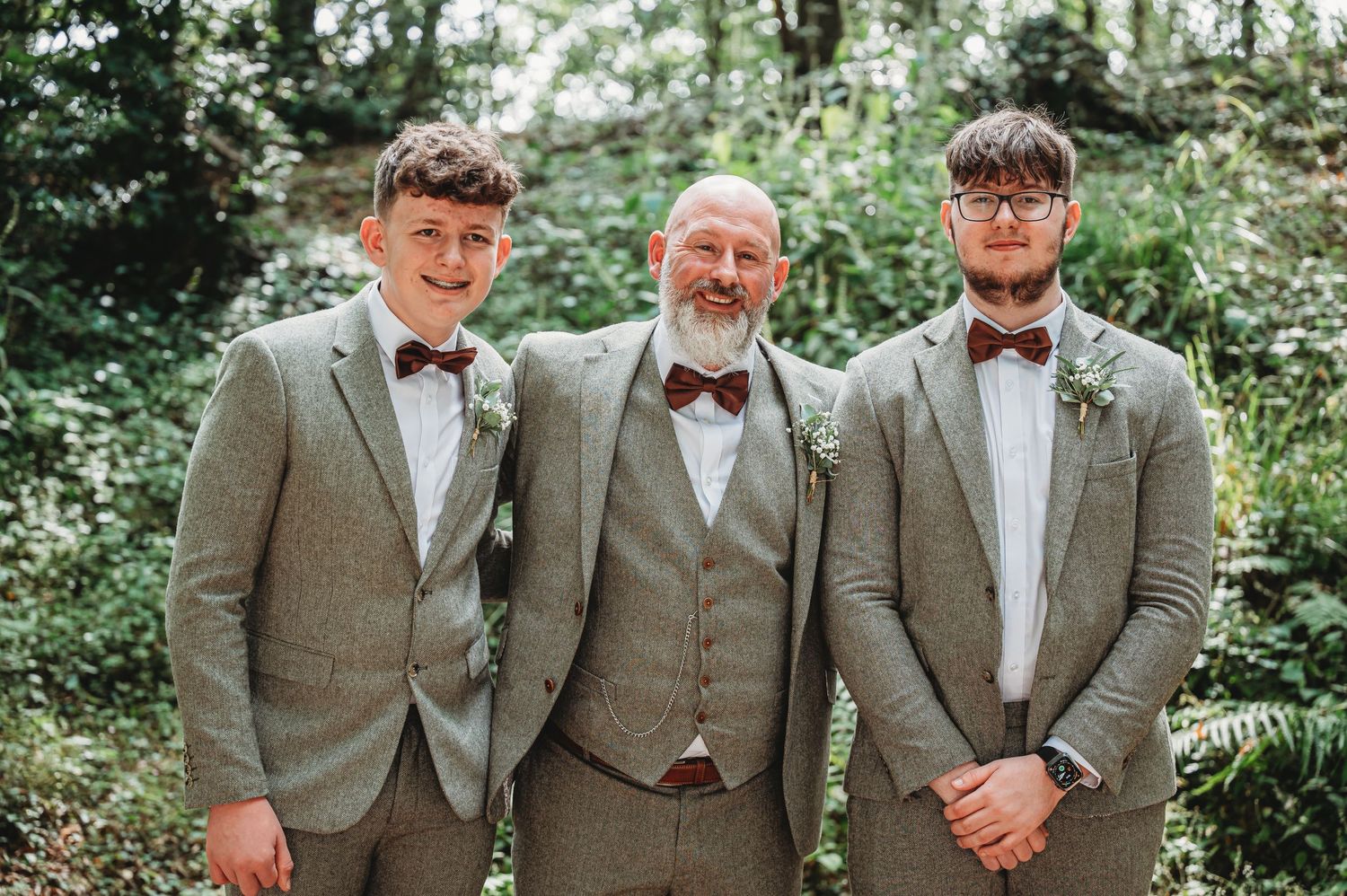 Three people in matching grey suits and bow ties pose together at an outdoor wedding venue surrounded by greenery.