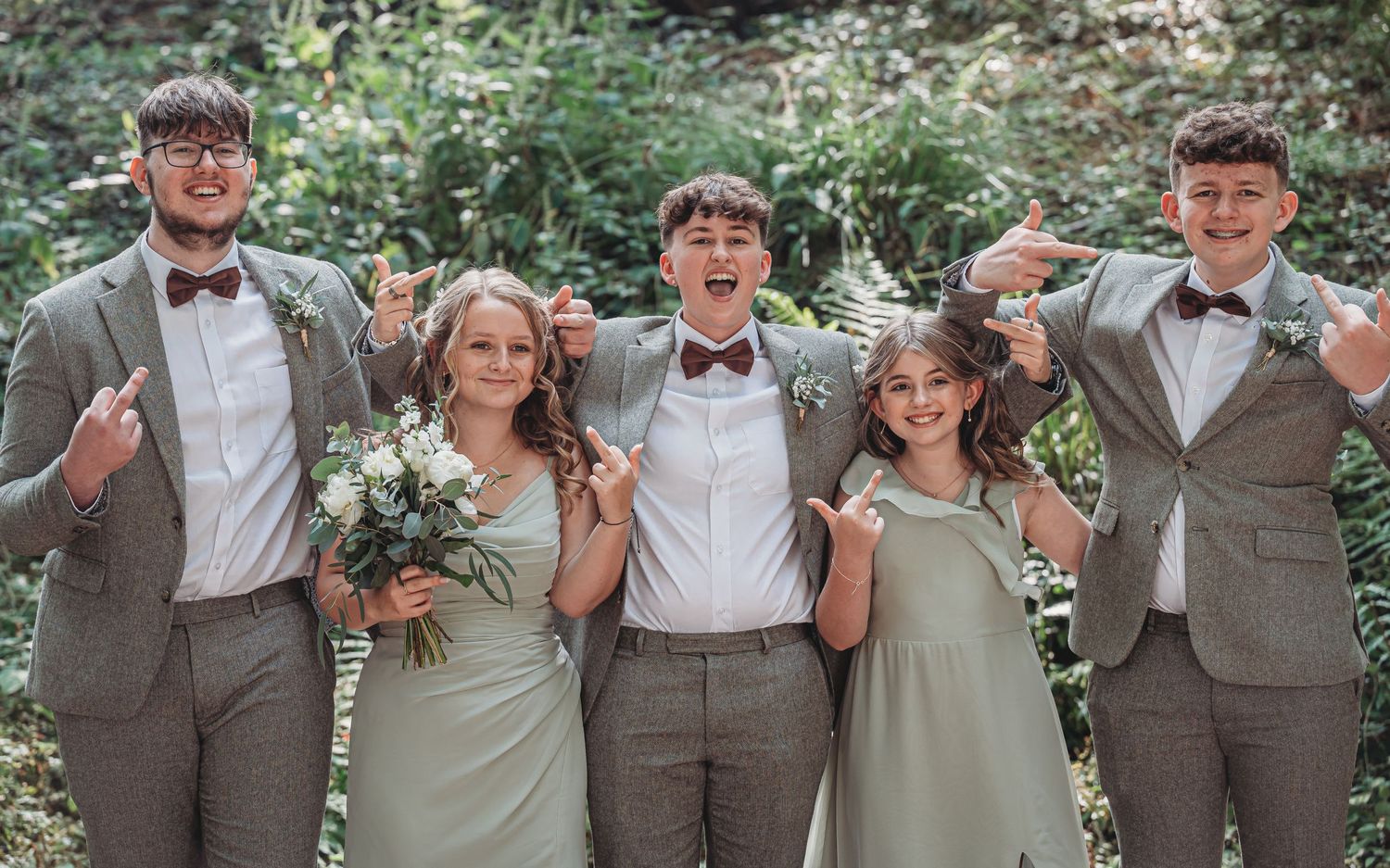 Wedding party in coordinated gray suits and sage green dresses poses together outdoors in a garden setting.