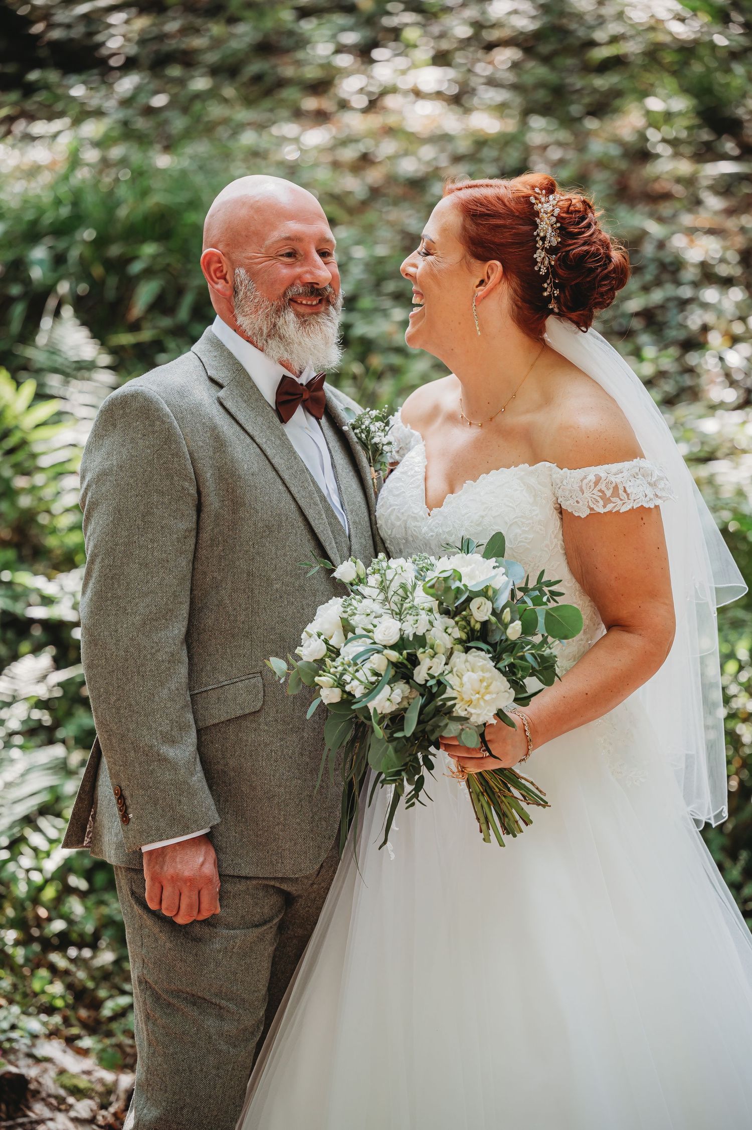 A joyful wedding couple sharing a laugh in a garden setting while holding an elegant white and green bouquet.