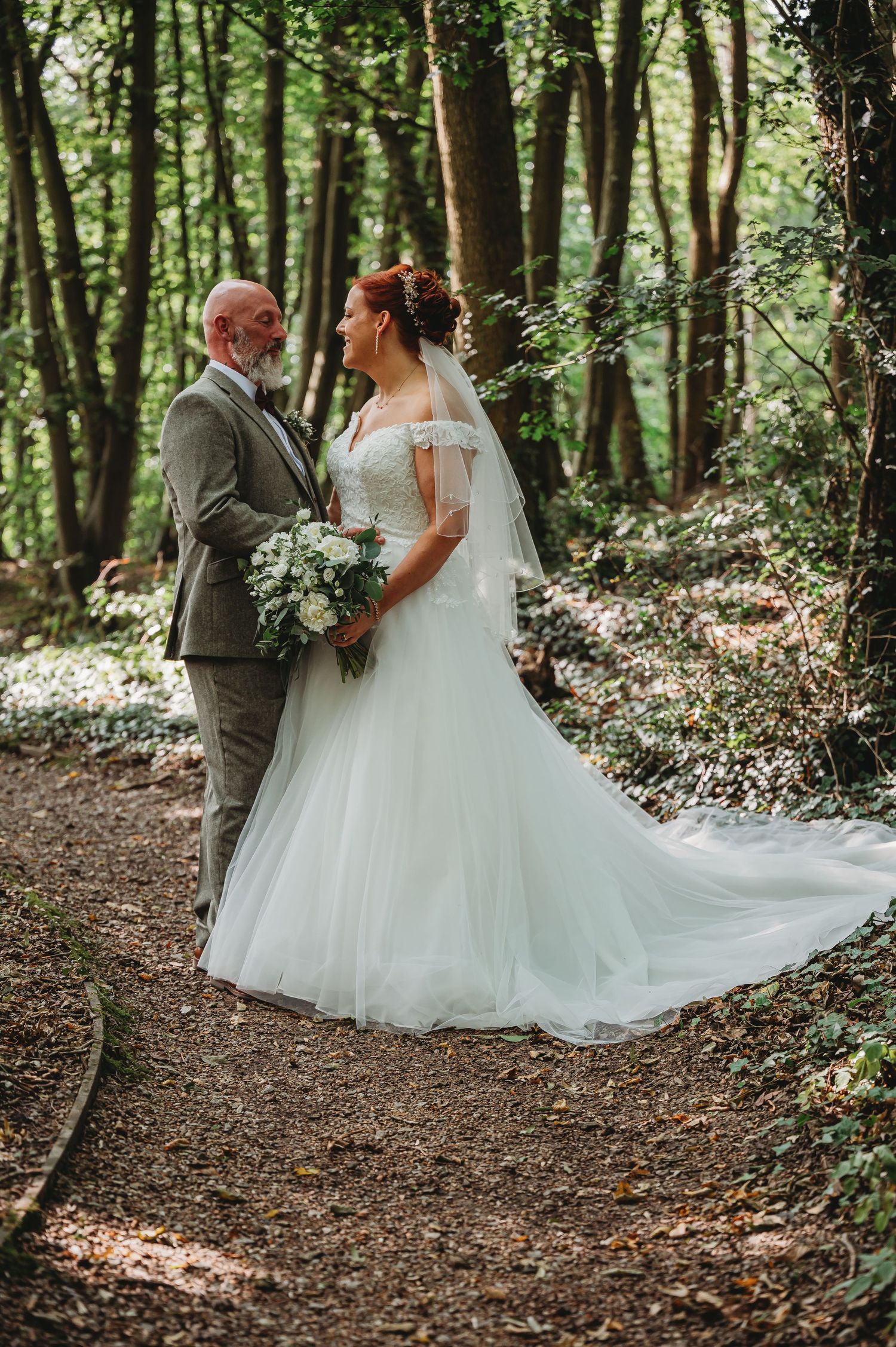 A romantic woodland wedding photo captures the bride in a flowing white gown and groom in gray suit on a forest path.