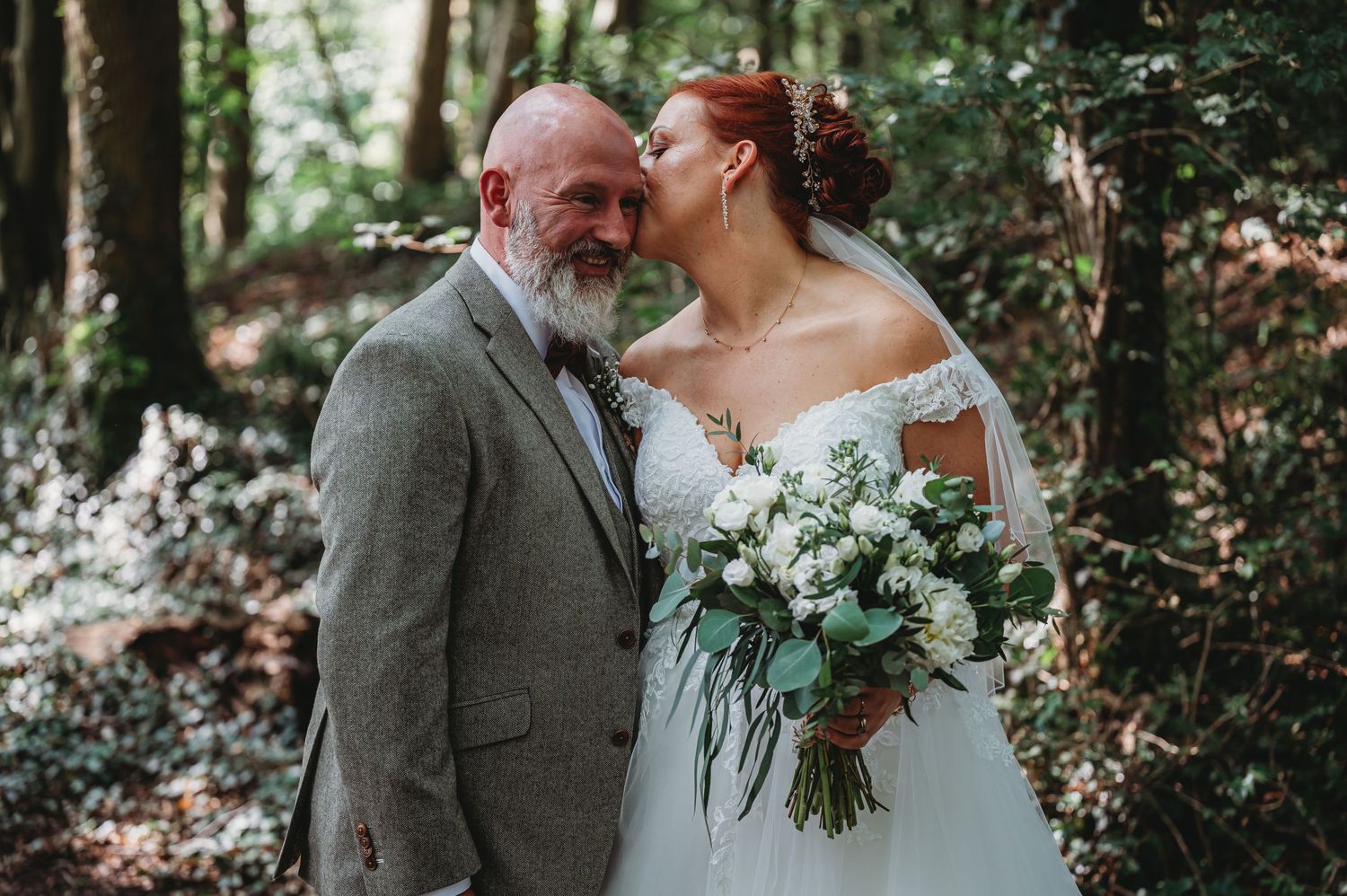 A couple shares an intimate moment in a forest setting during their wedding ceremony, with white floral bouquet in hand.