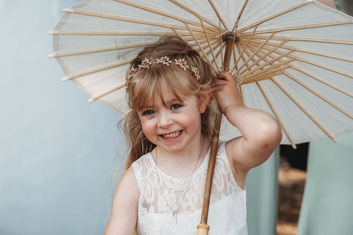 A young child in a white lace dress smiles while holding a vintage paper parasol against a light blue background.