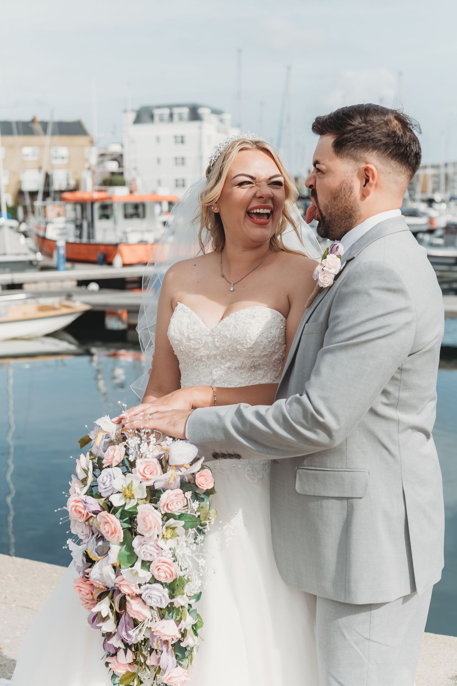 A joyful wedding couple shares a candid moment together at a marina with boats in background and cascading floral bouquet.