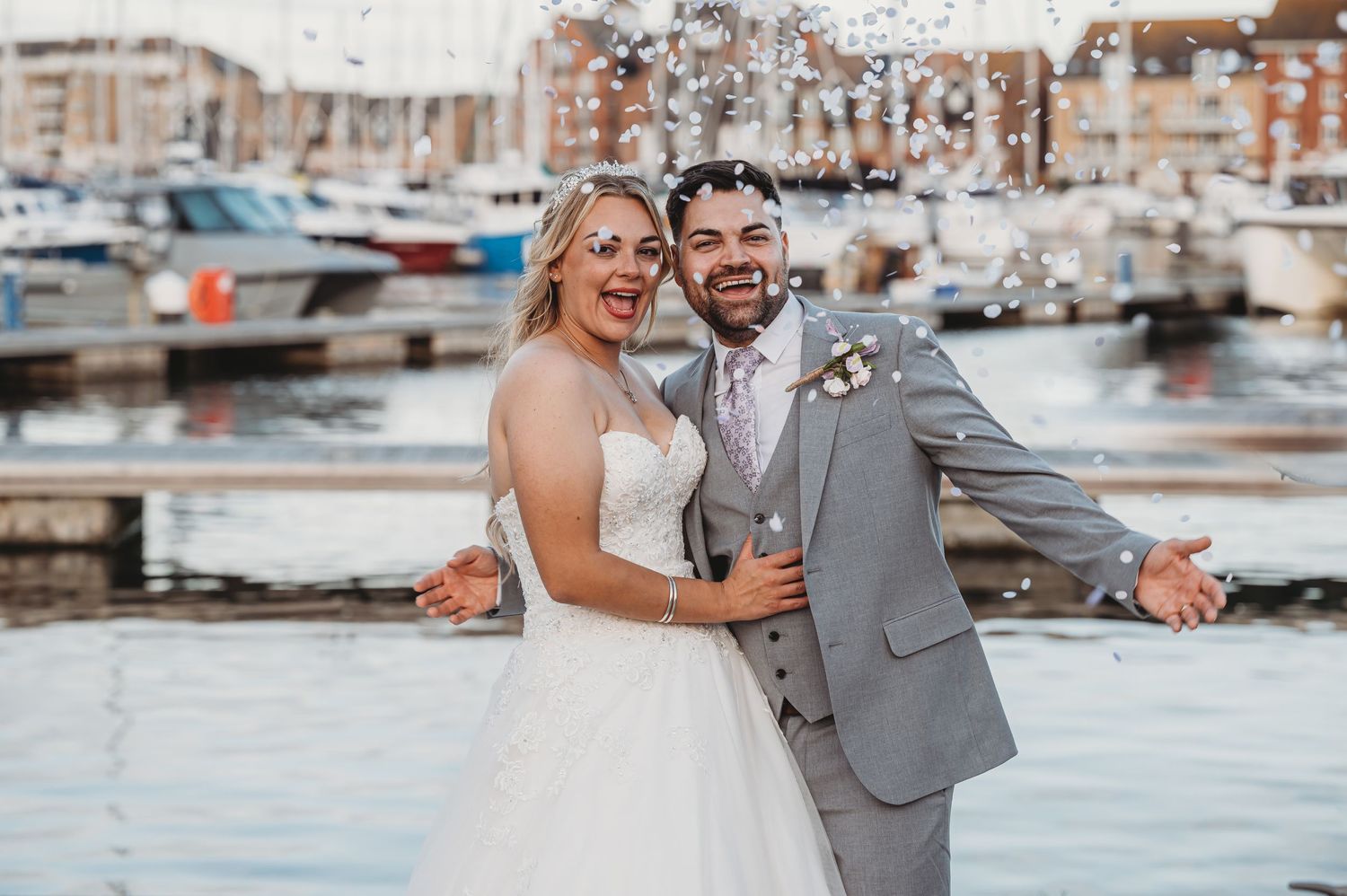 Newlyweds share a joyful moment together at a marina with boats and warm sunset lighting in the background.