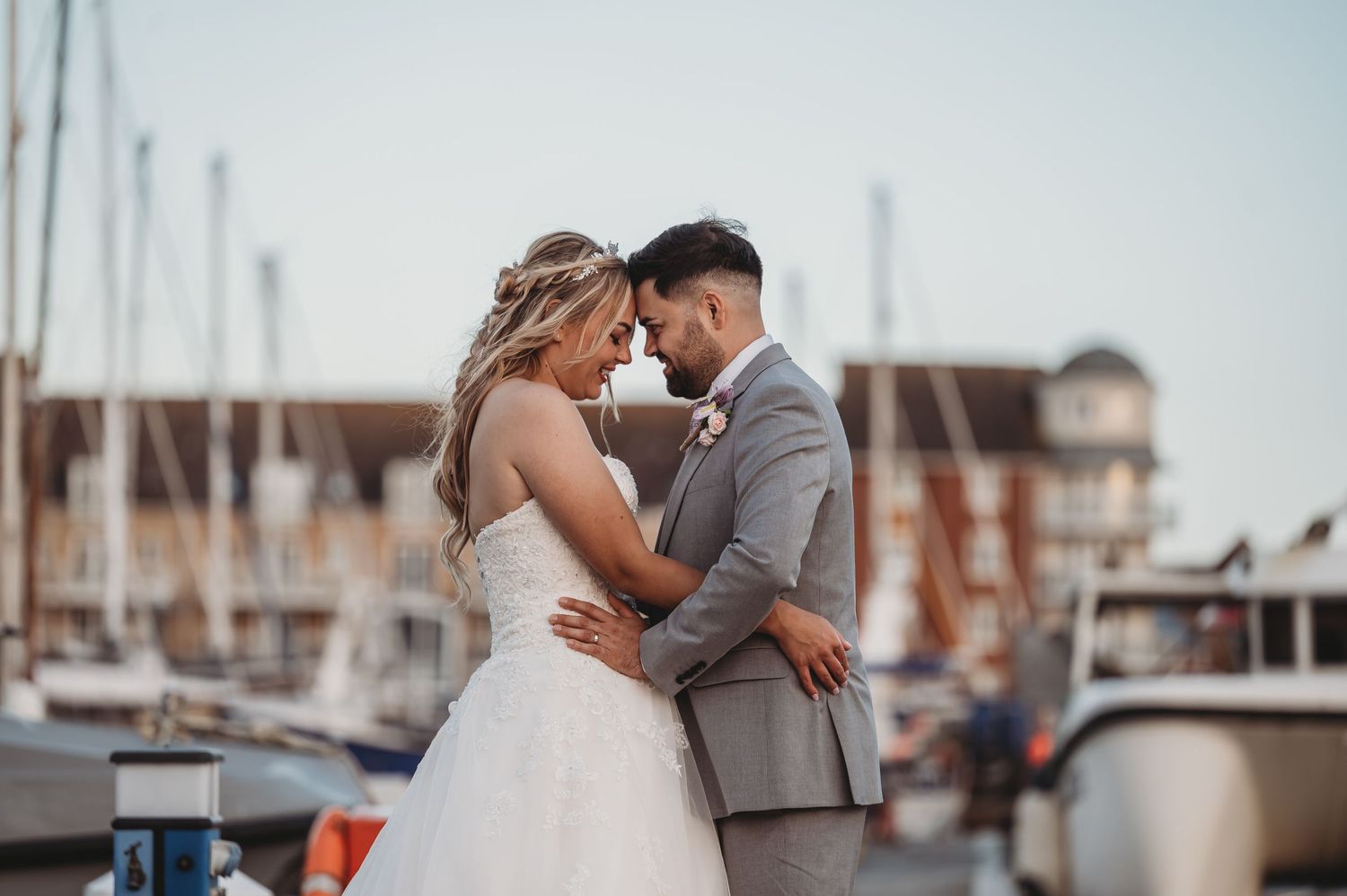 A romantic wedding couple embraces on a marina dock at sunset with boats and harbor in the background.