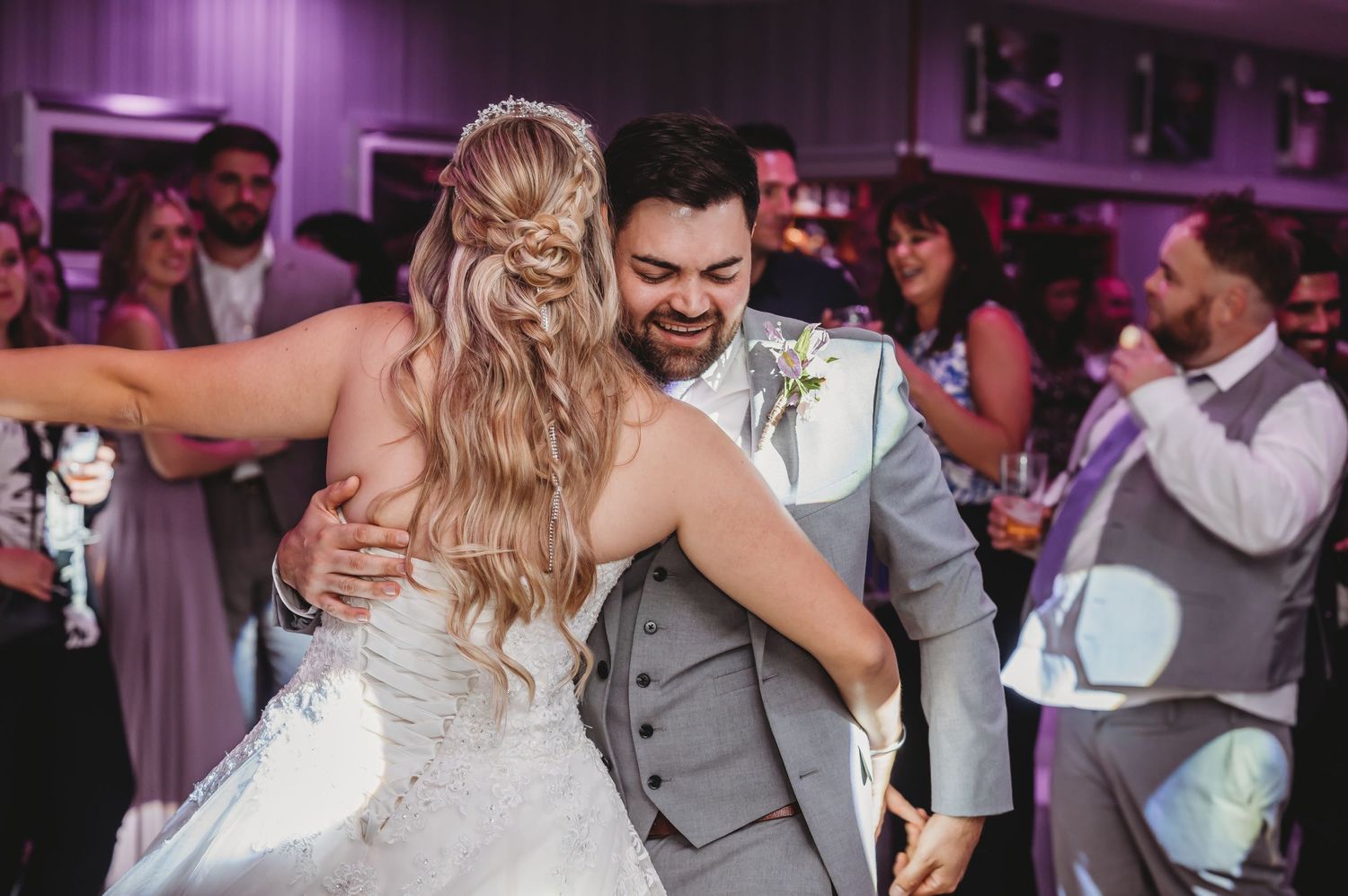 A couple shares their first dance at a wedding reception with purple lighting and guests watching.