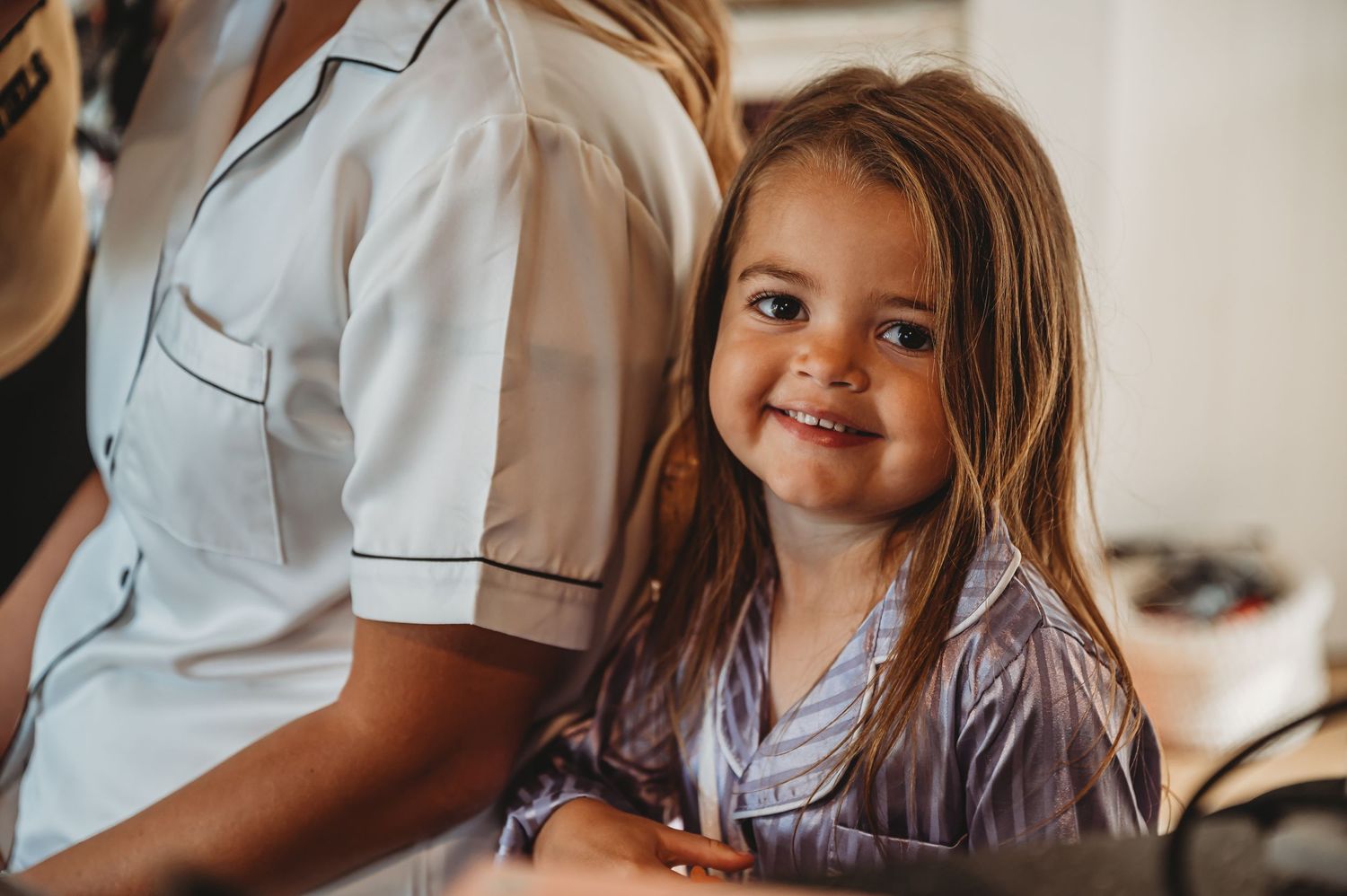 Child with long brown hair in braids wearing a lavender shirt smiles warmly while sitting indoors.