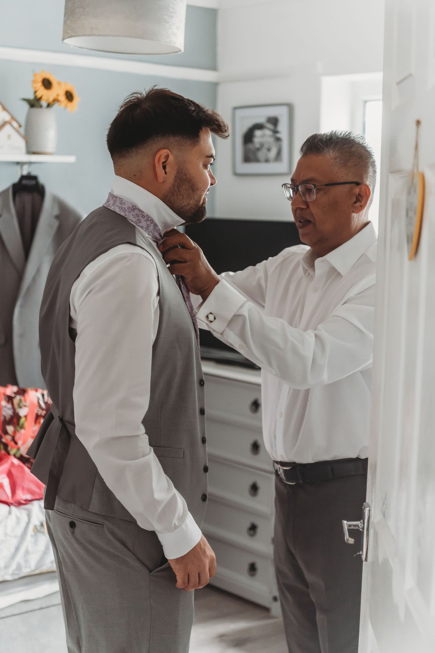 Two people in a bedroom getting dressed for a formal occasion, with one adjusting the other's tie.