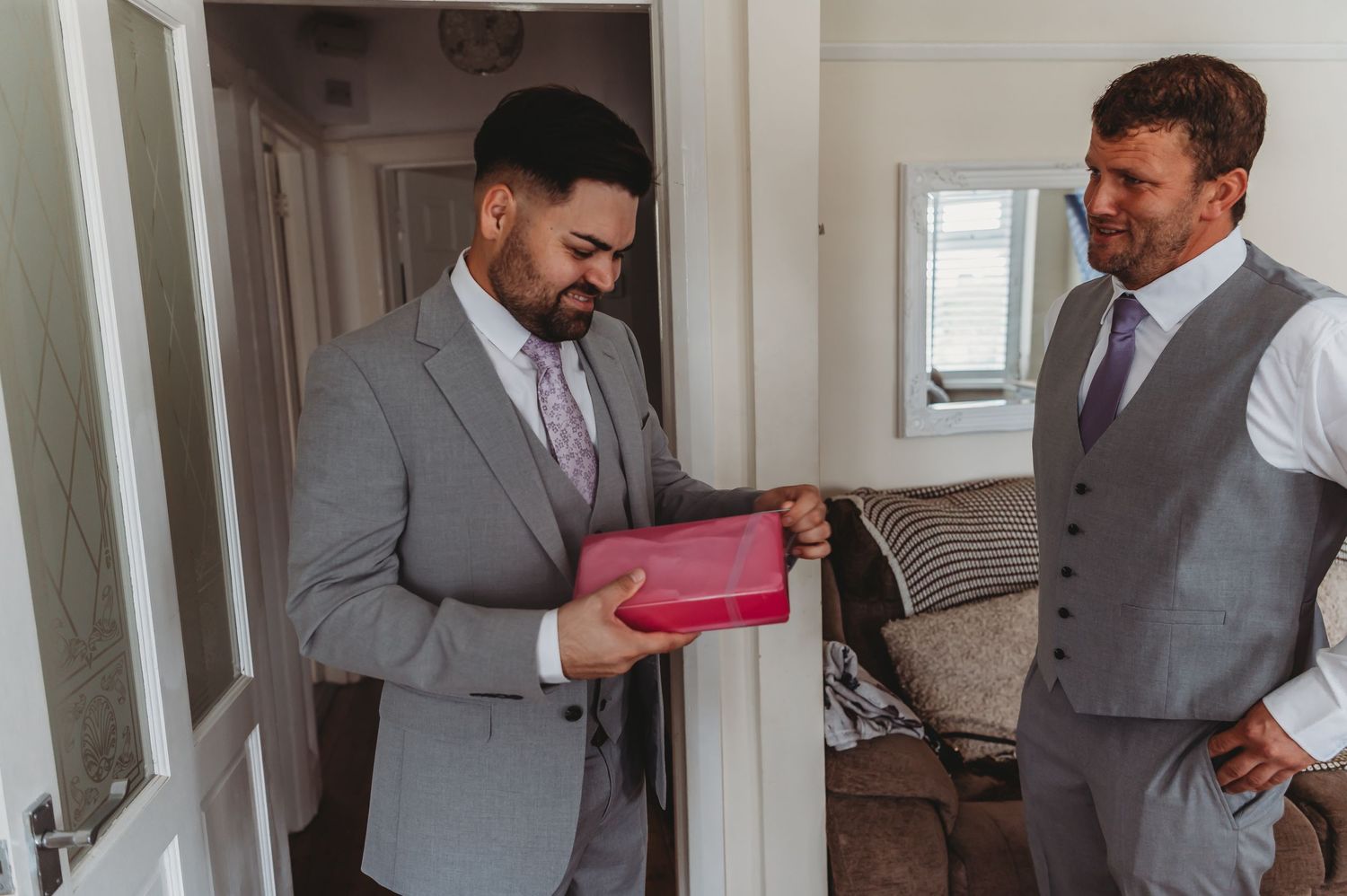 A man in a gray suit smiles while opening a pink gift box as another looks on in a bright indoor space.