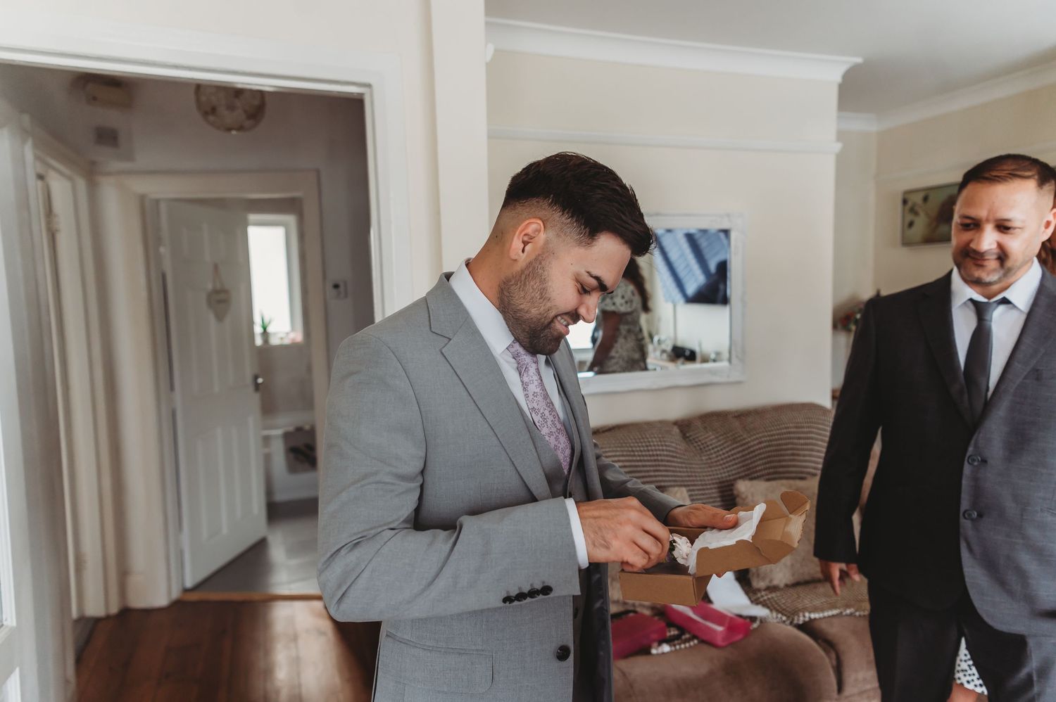 Two men in formal suits share a moment of joy while examining a small pink package in a bright indoor setting.