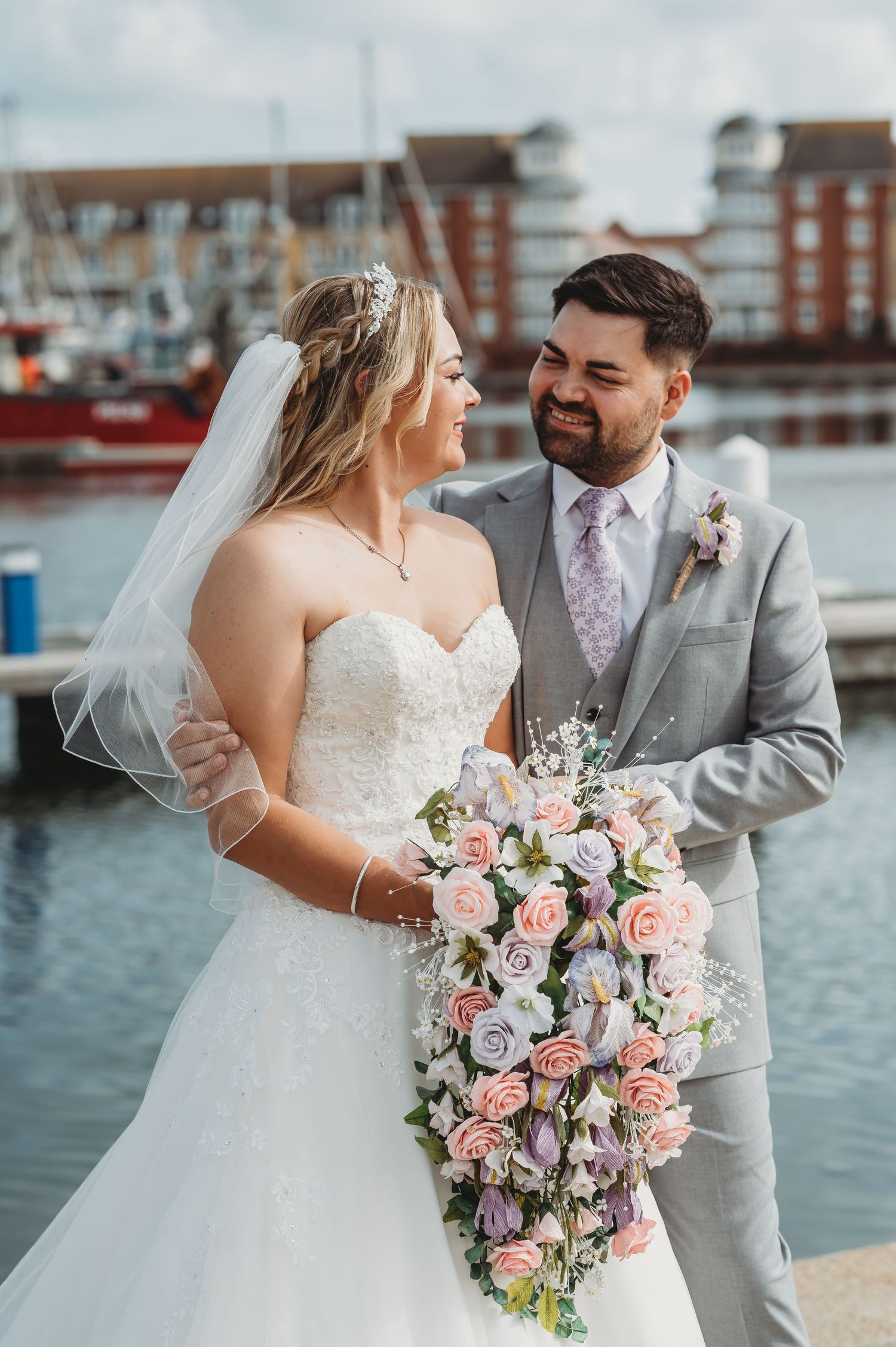 Newlywed couple share tender moment by marina with cascading pink and purple floral bouquet.