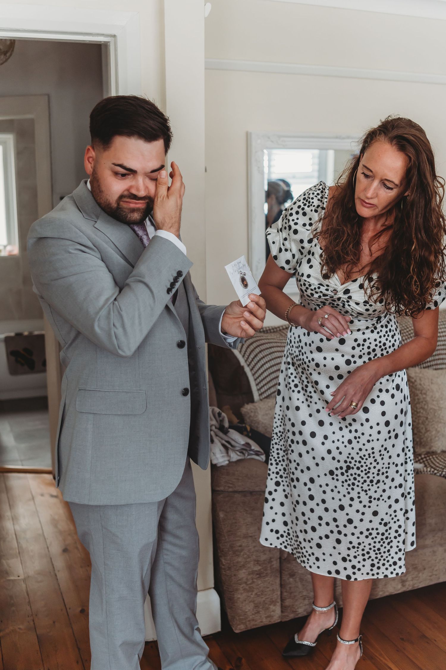 Two people in formal attire stand in a home interior, one wearing a grey suit and one in a polka dot dress.