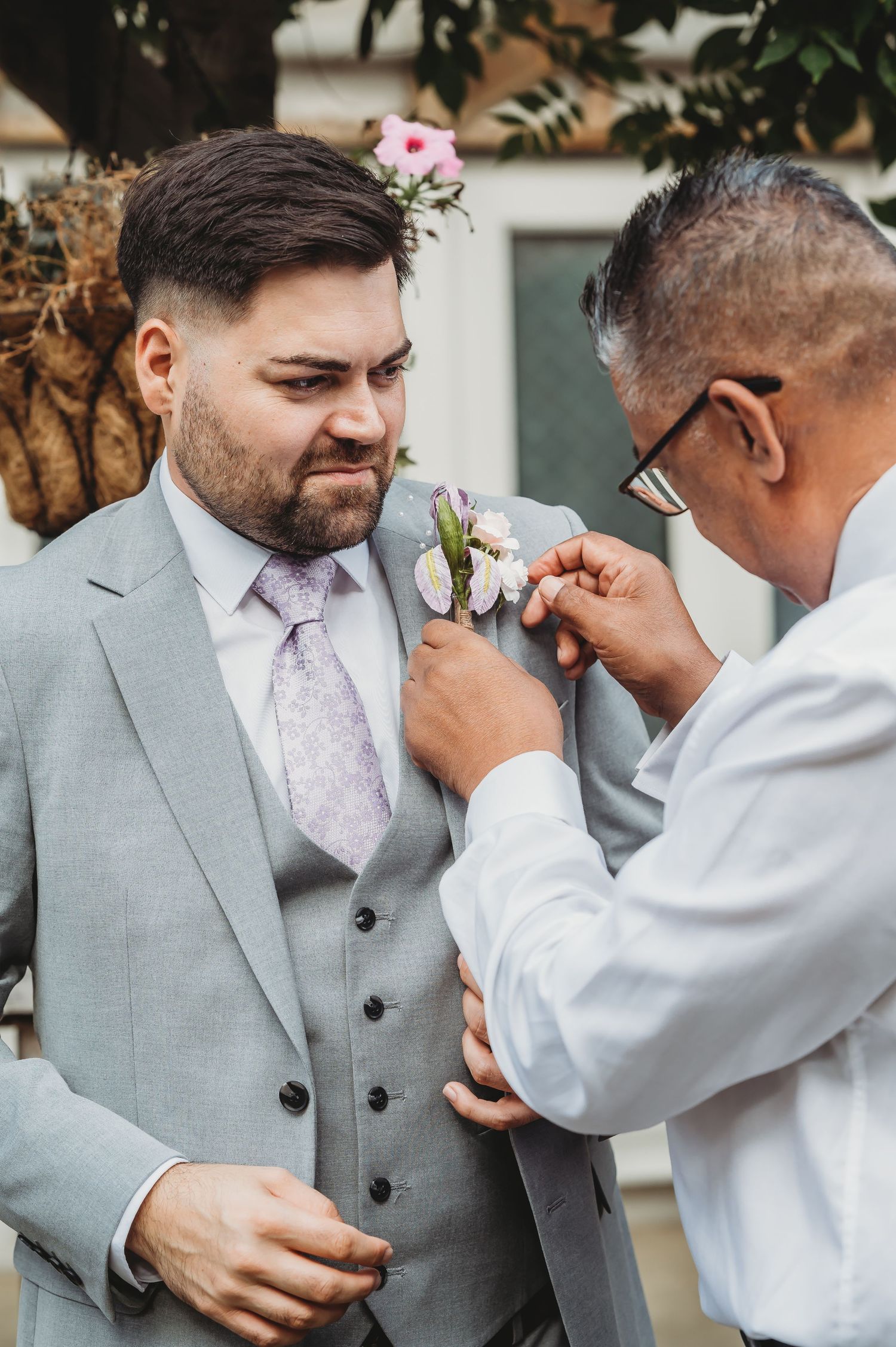 A groomsman helps adjust a white floral boutonniere on a grey three-piece suit at an outdoor wedding ceremony.