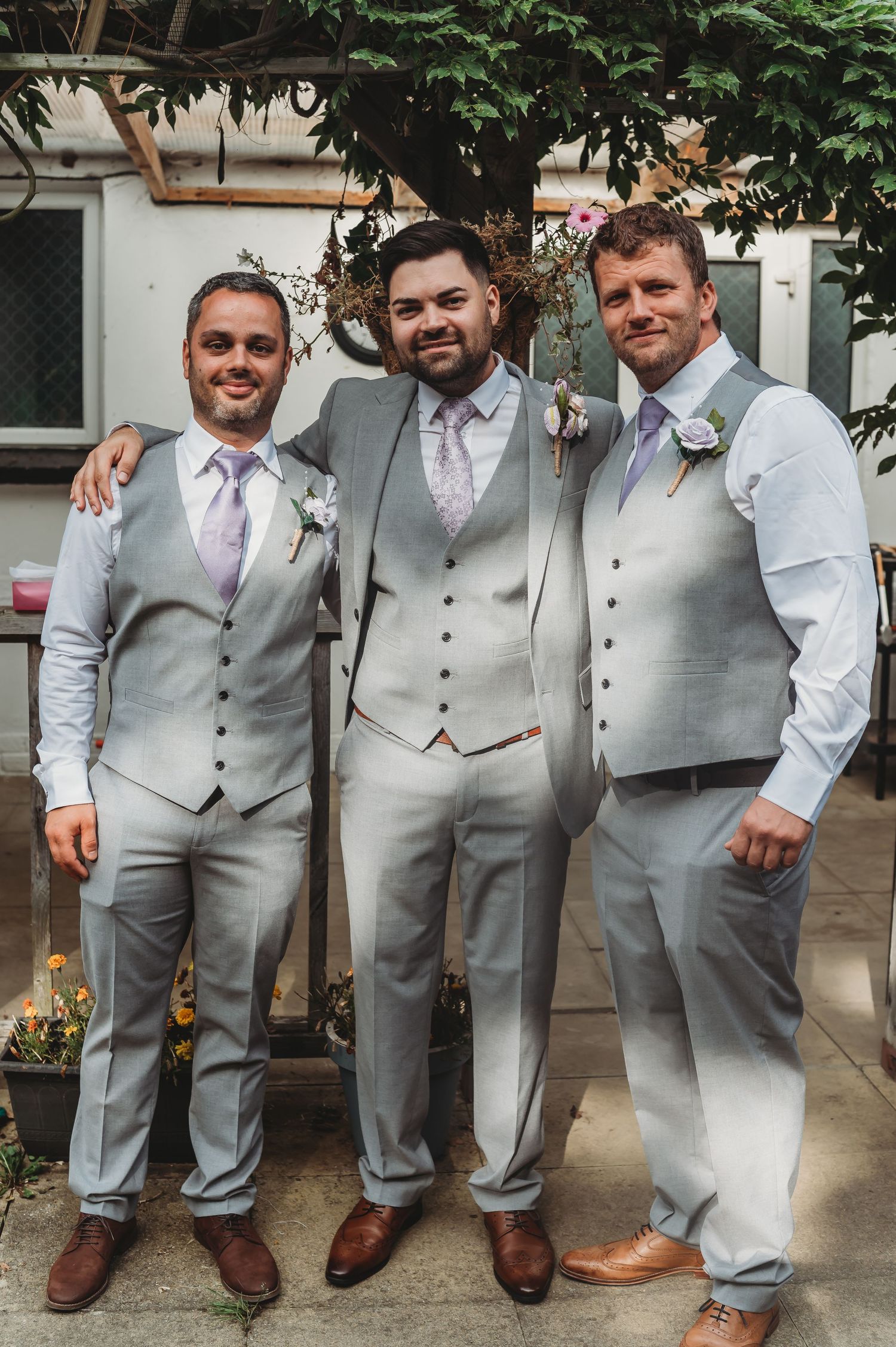 Three groomsmen pose together in matching light gray vests and suits with lavender ties for a wedding photo.