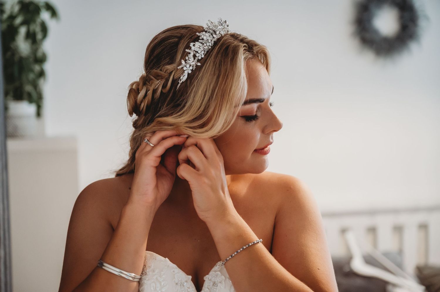 Side profile of a bride wearing a delicate tiara and strapless dress while touching her ear on her wedding day.