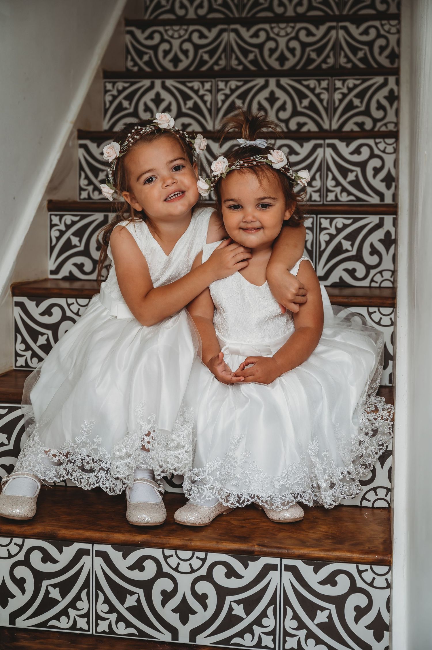 Two little girls in white lace dresses and floral headpieces sit together on decorative tiled stairs.