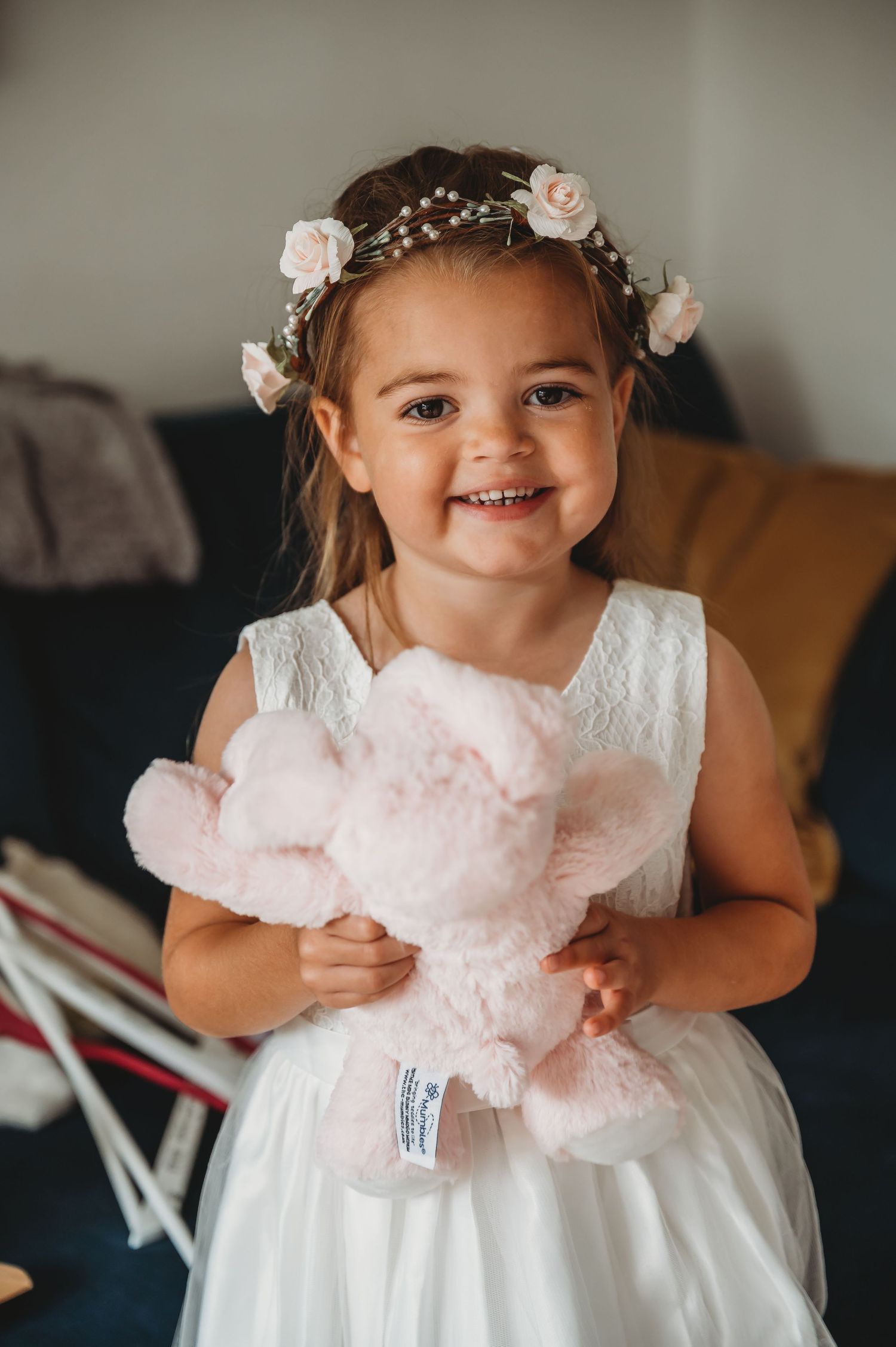 A child in a white dress and floral crown smiles while holding a pink plush toy.