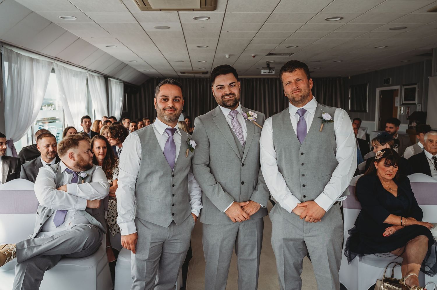 Three groomsmen in matching light gray vests and ties stand together at an indoor wedding ceremony venue.