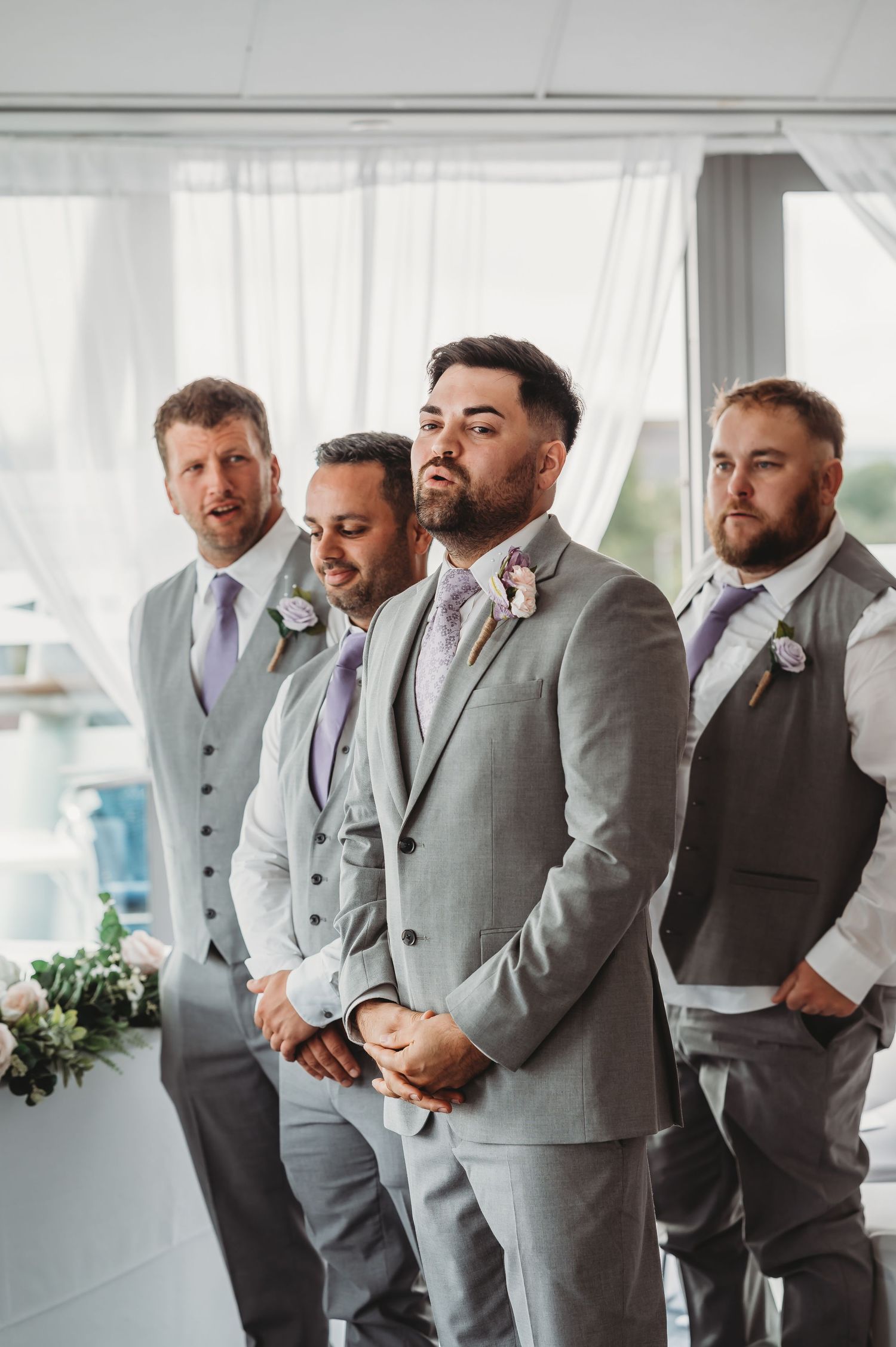 Four groomsmen in coordinated gray suits and vests stand together at an elegant indoor wedding venue.