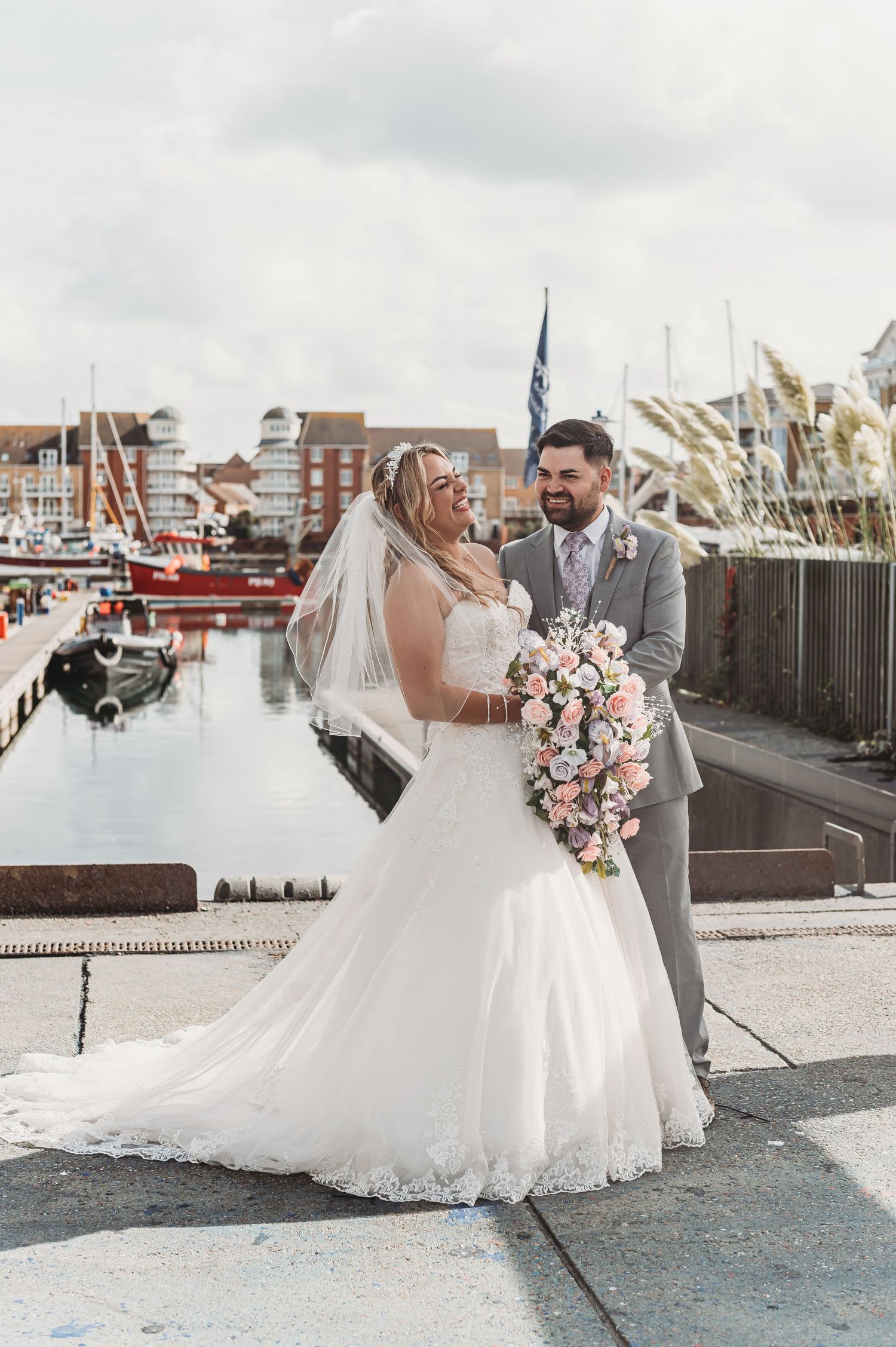 A romantic wedding couple shares a tender moment at a marina with boats and buildings in the background.