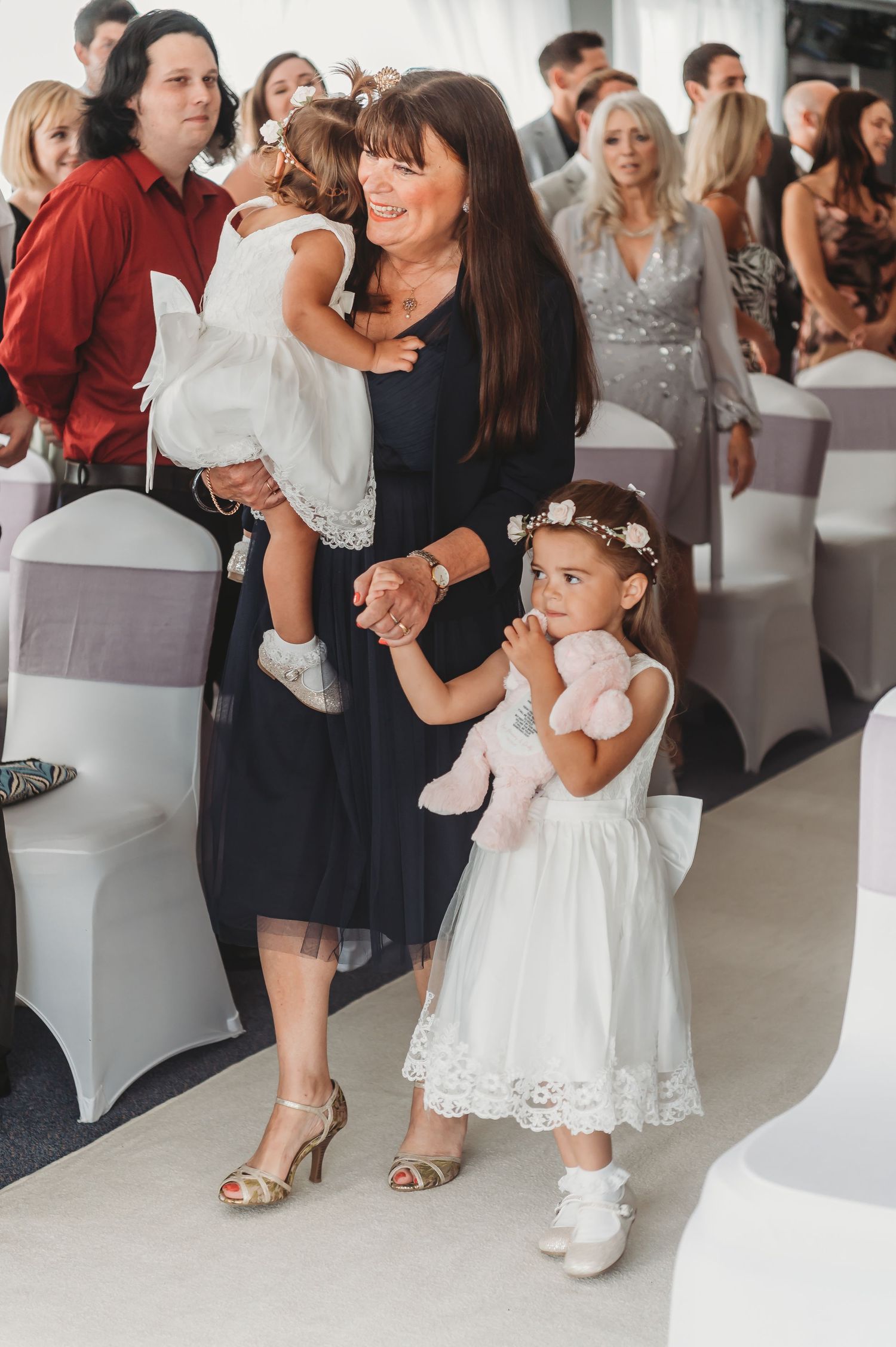 A woman in a navy dress stands with two young girls in white dresses at a formal event with covered chairs.