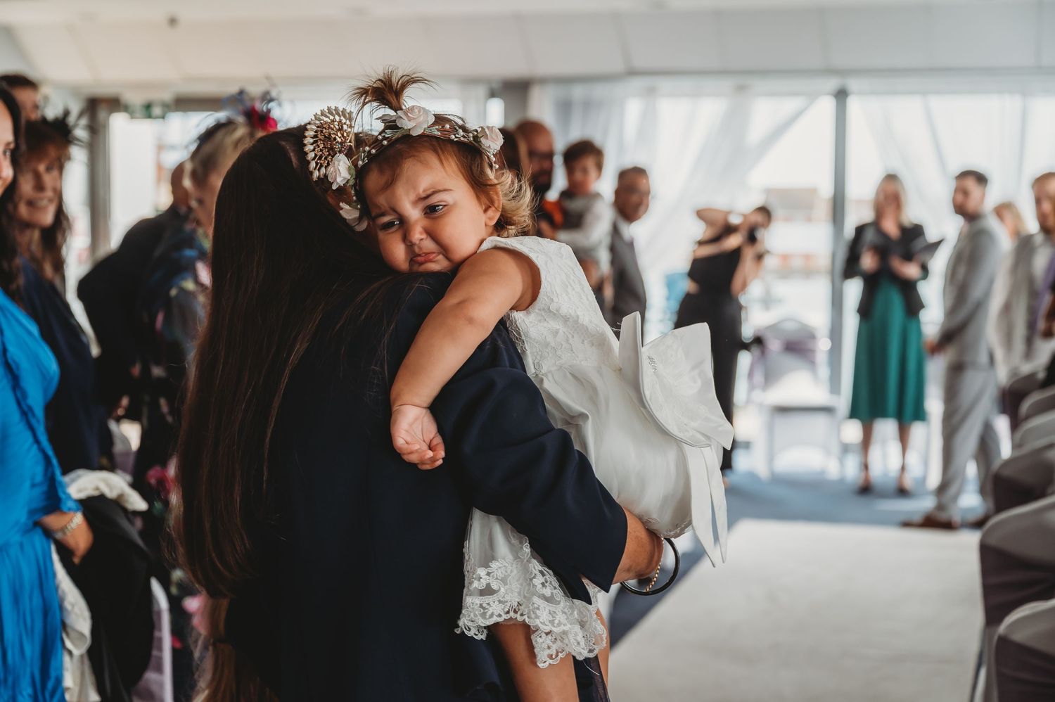A sleepy toddler in a white dress rests on someone's shoulder during a formal gathering with guests in background.