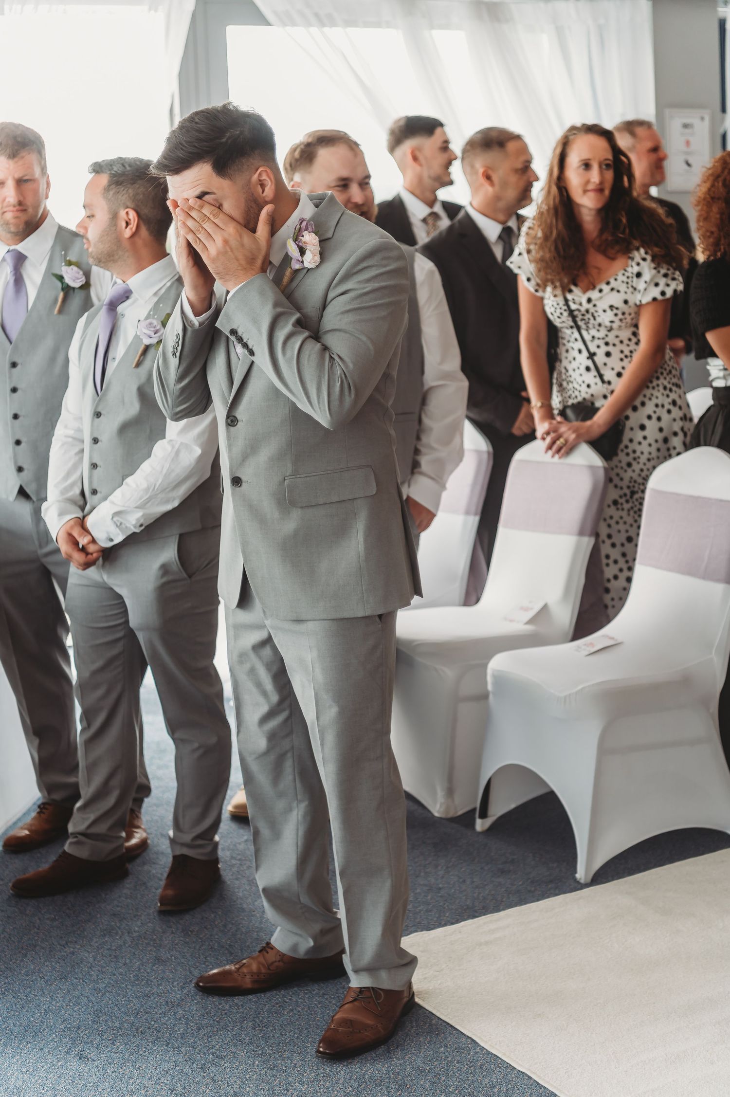Emotional groomsman covers his face during wedding ceremony while standing with other groomsmen in gray suits.