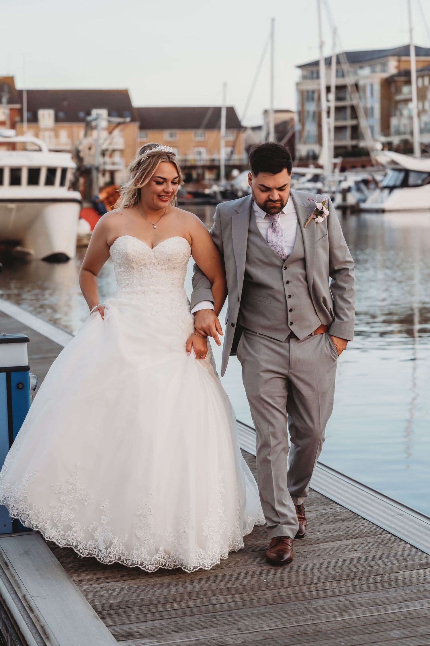Romantic wedding photo on marina boardwalk with boats and buildings in background.