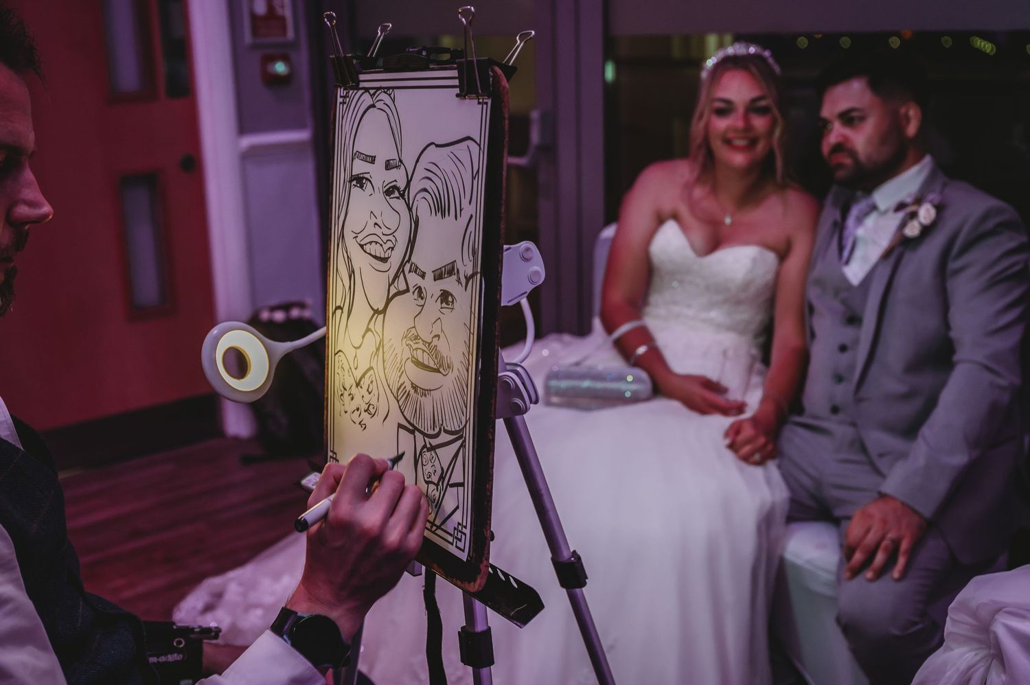 An artist sketches a caricature on a mounted easel while two wedding guests sit nearby during the reception.