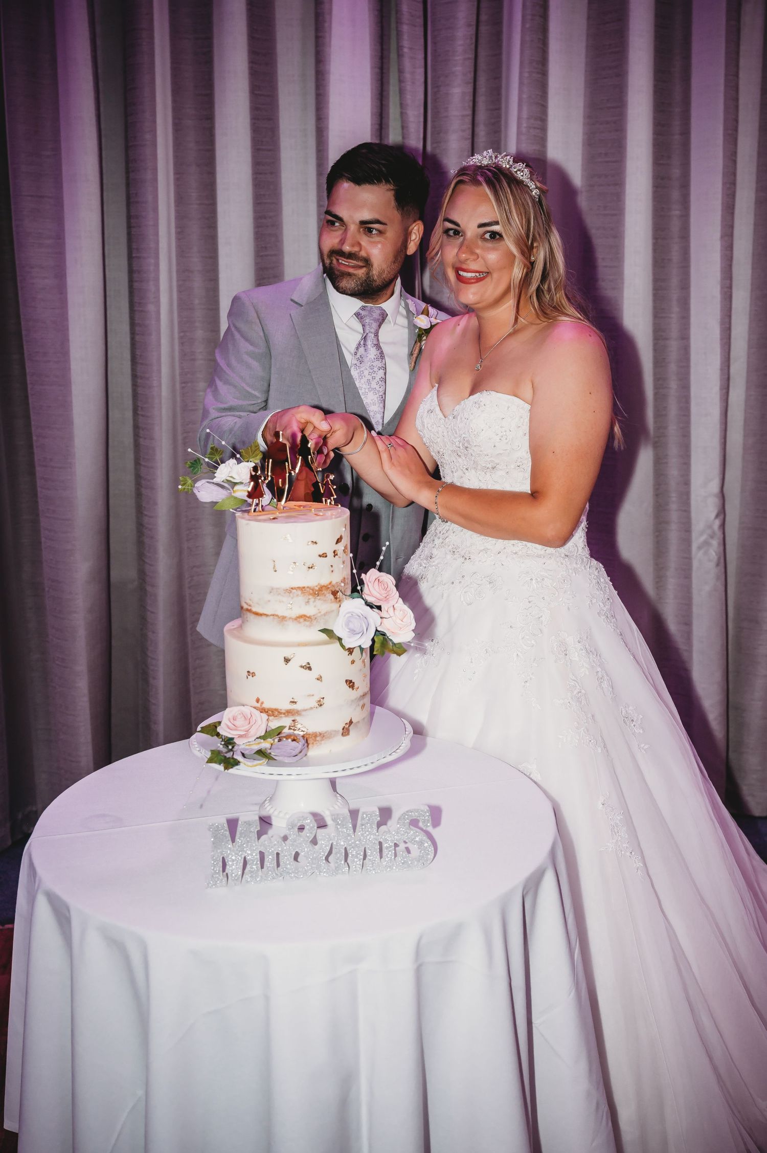 A bride and groom stand next to their elegant three-tiered wedding cake at their reception with purple lighting.