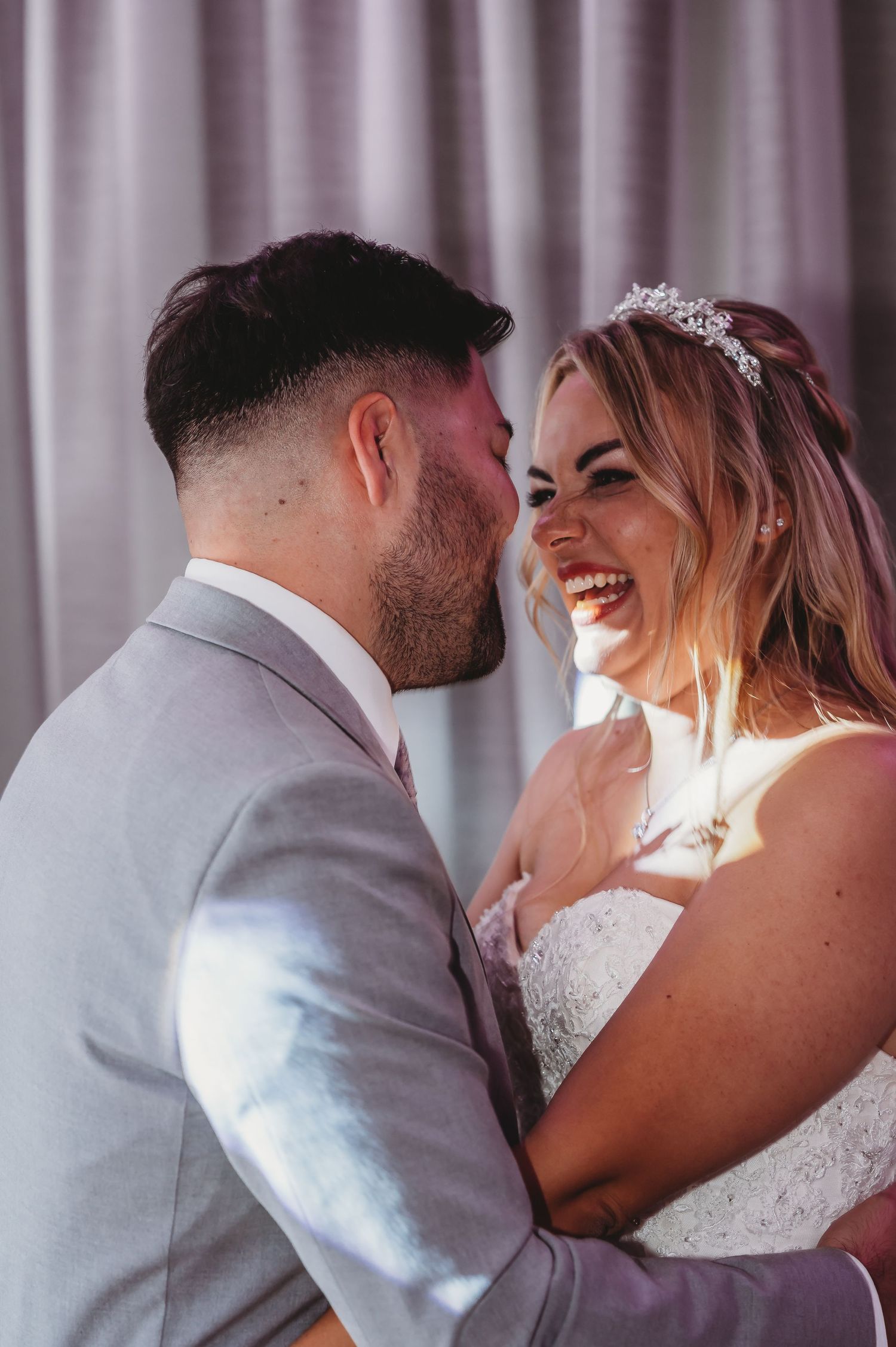 Romantic candid moment between newlyweds against light purple curtain backdrop.