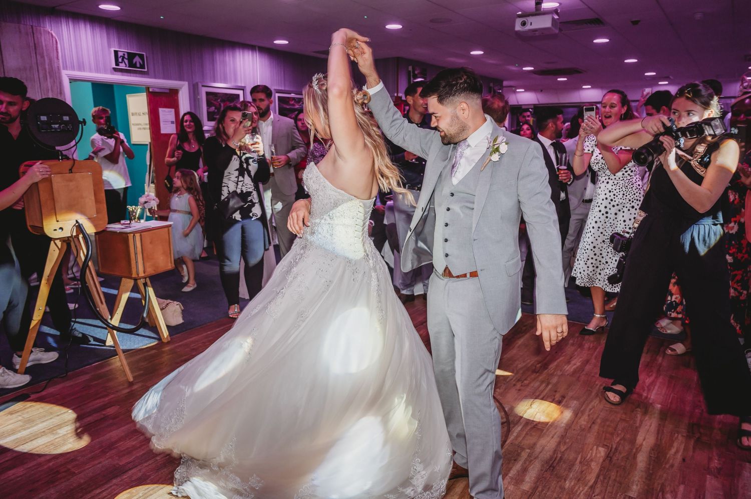 A couple shares their first dance in a purple-lit reception venue while guests watch and take photos on the dance floor.