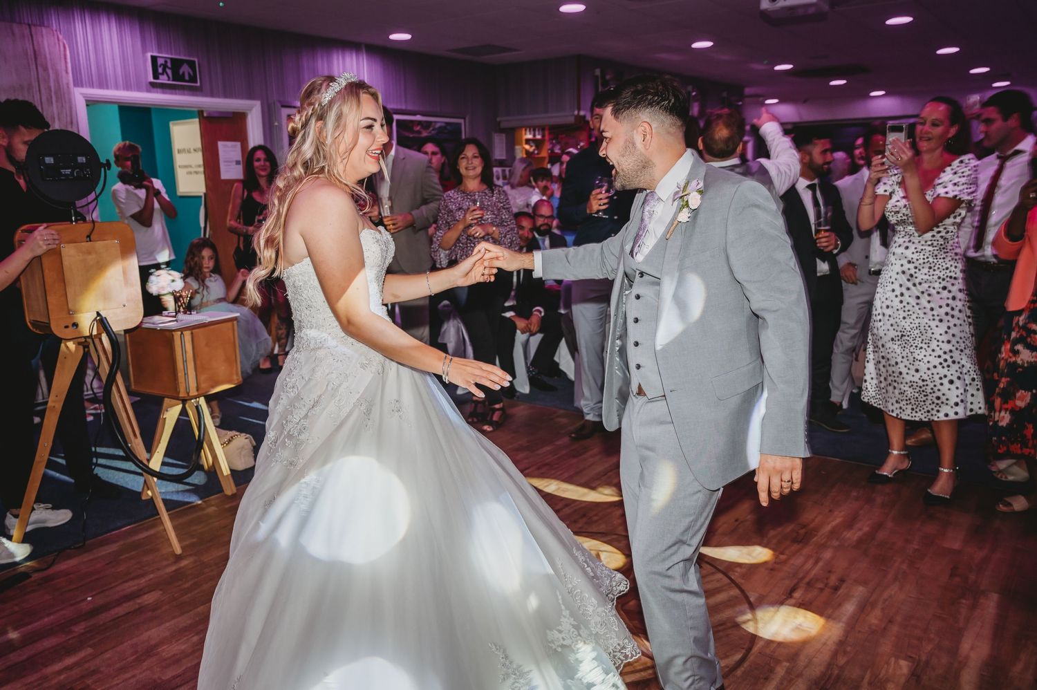 Newlywed couple shares their first dance on a wooden floor at their wedding reception with purple lighting and guests watching.