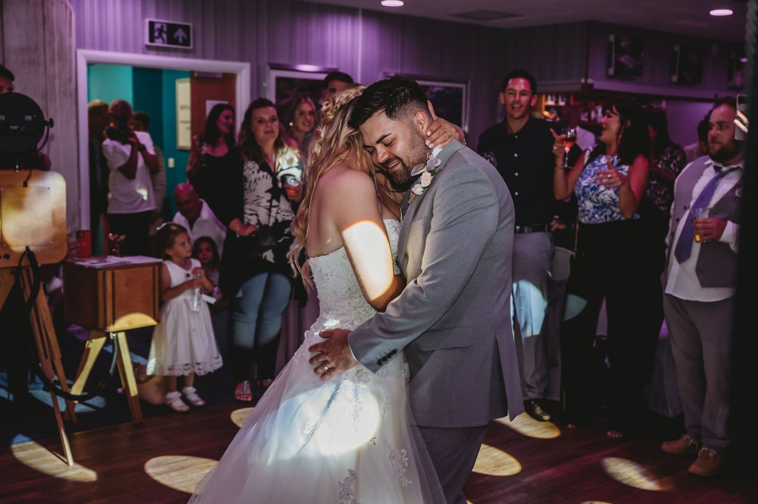 A couple shares their first dance at their wedding reception as guests watch from the edges of a purple-lit dance floor.