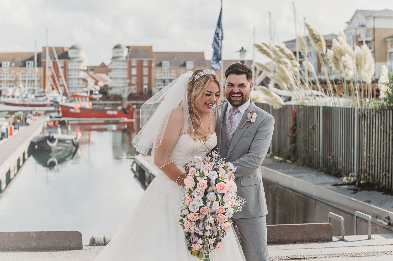 A happy wedding couple posing at a marina with boats and apartments in the background.