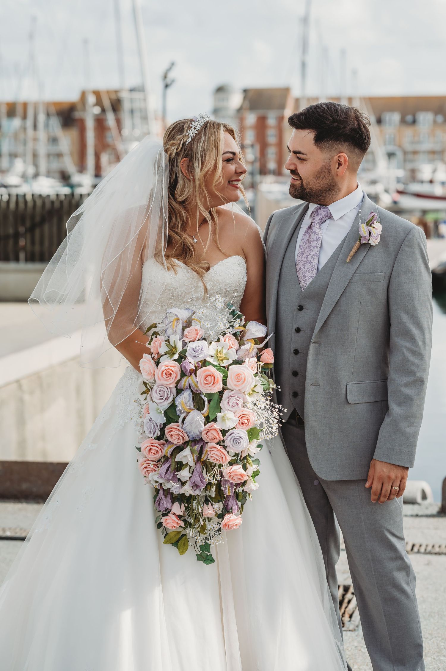 A newlywed couple stands at a marina, with the bride holding a cascading bouquet of pink and white flowers.