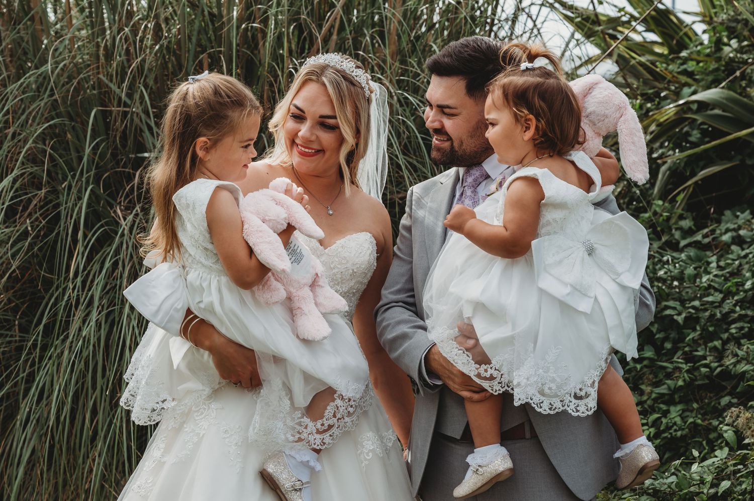 Outdoor wedding portrait of bride and groom in formal attire holding two young children in white dresses against tall grass.