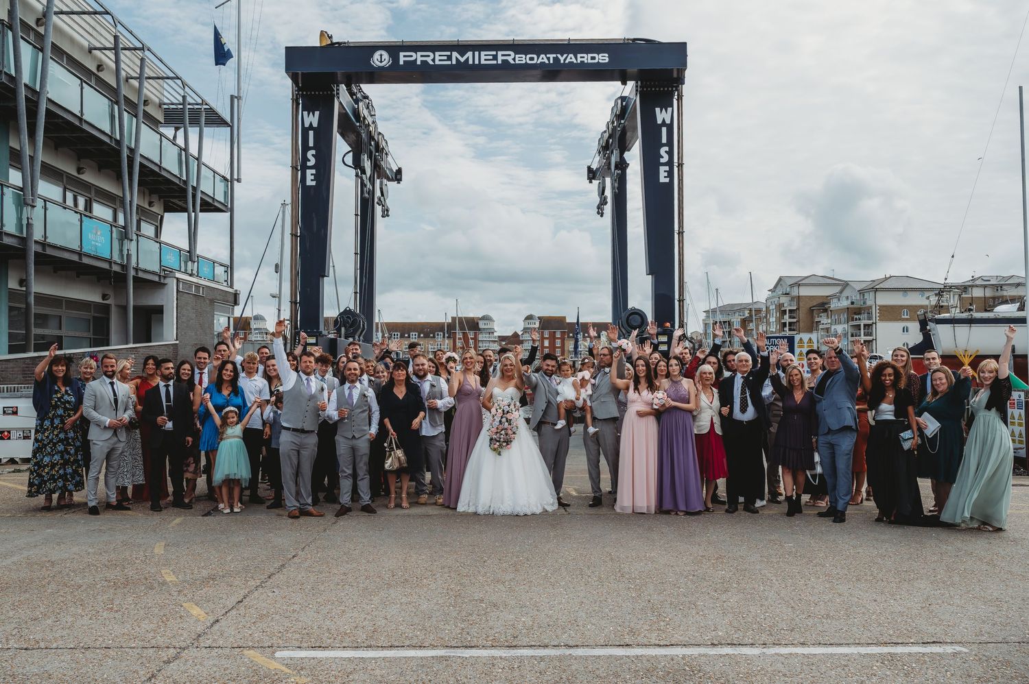 Large wedding party gathers for group photo beneath industrial crane at marina dock on cloudy day.