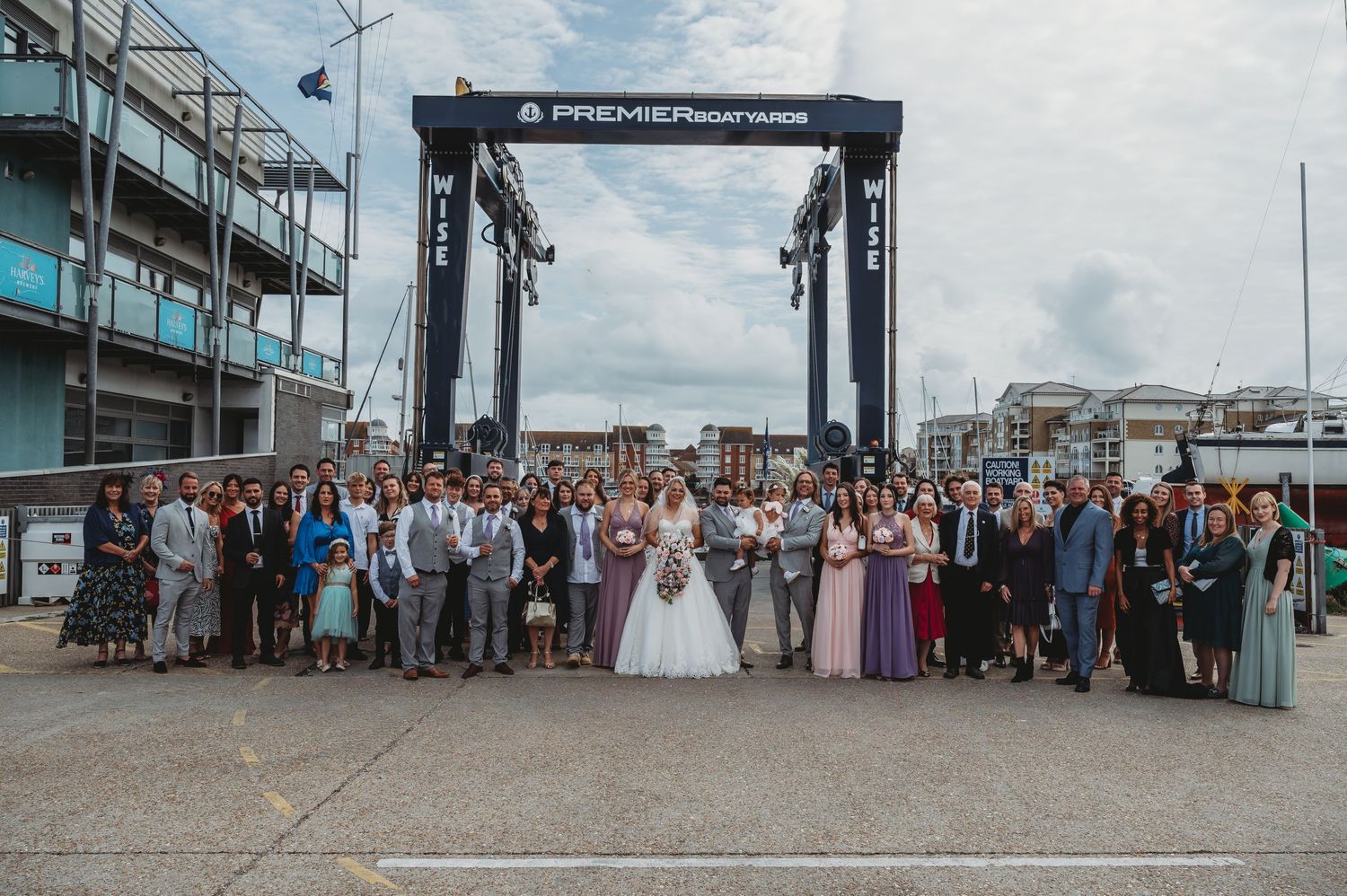 Large group wedding photo of guests and wedding party in front of an industrial marina backdrop.