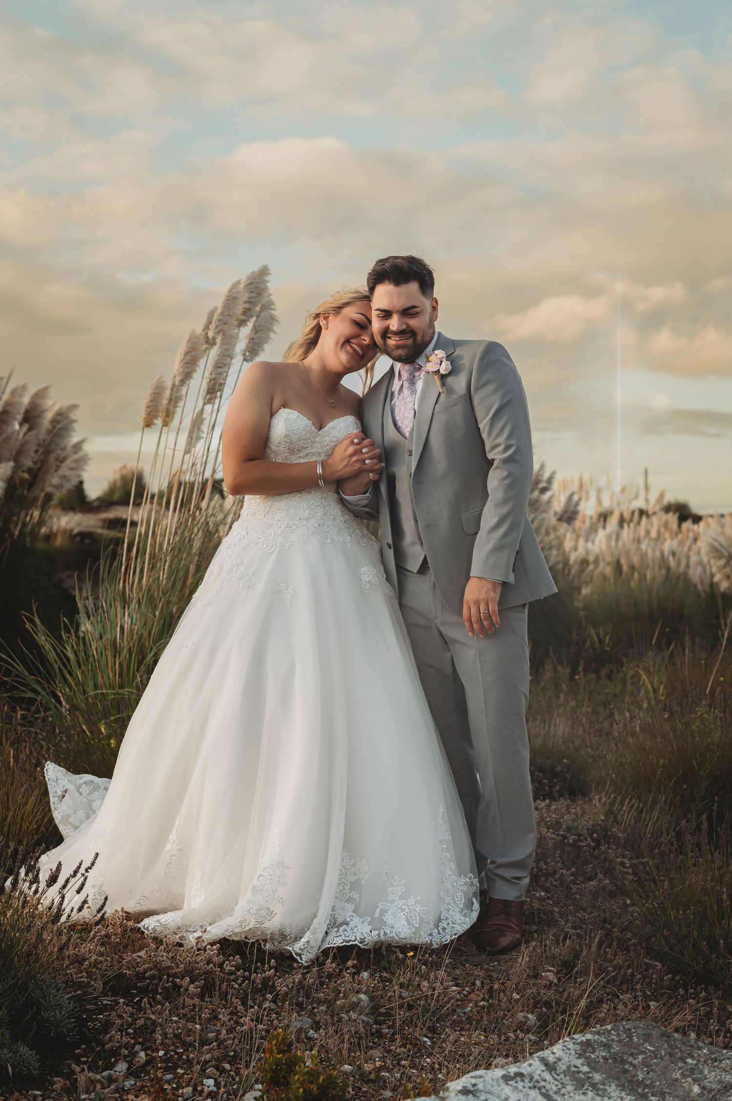 A couple in wedding attire poses together in front of tall pampas grass during golden hour at sunset.