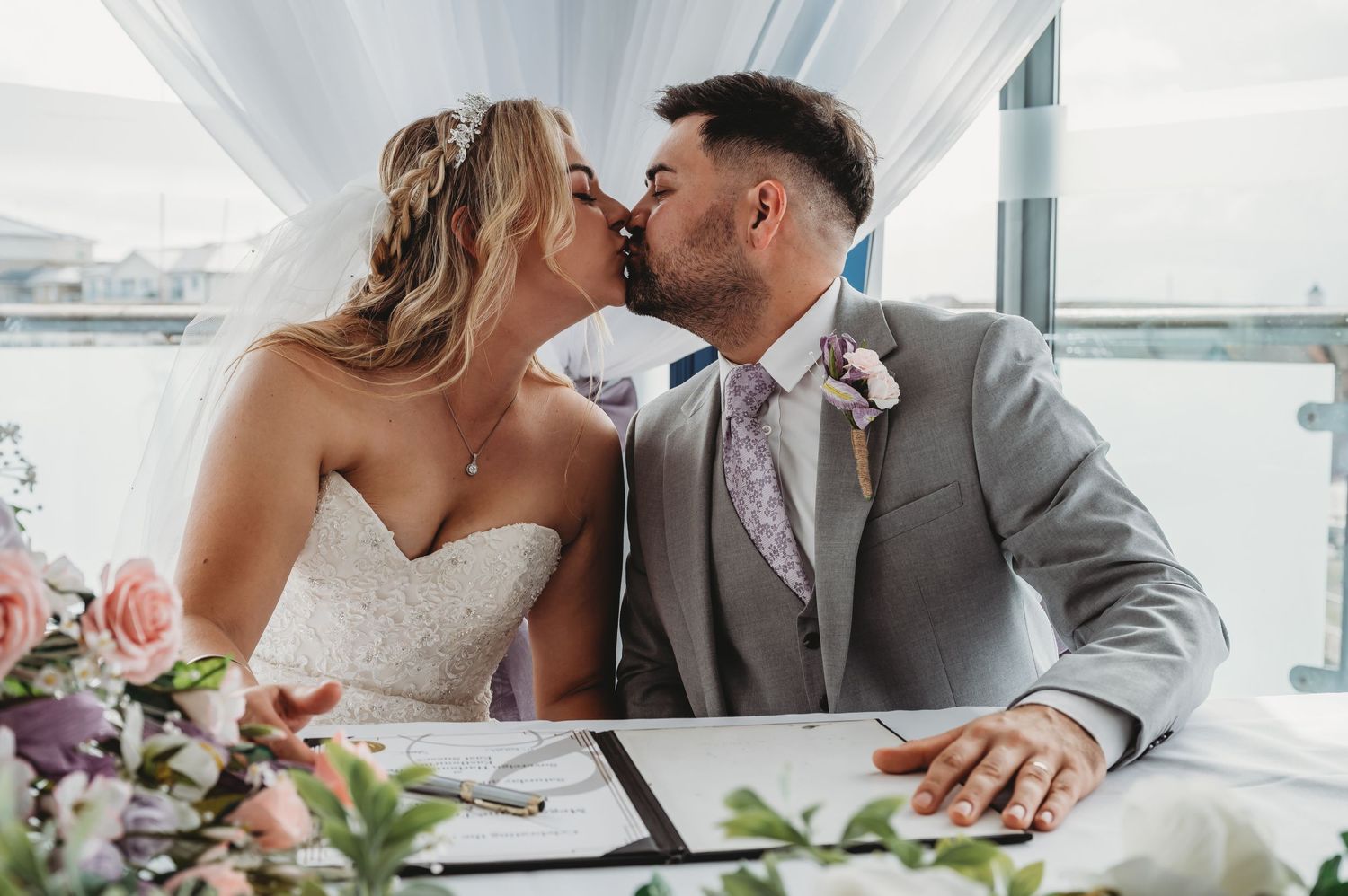 Newlyweds share a kiss while signing their marriage license at an elegant white-draped venue with pink floral arrangements.
