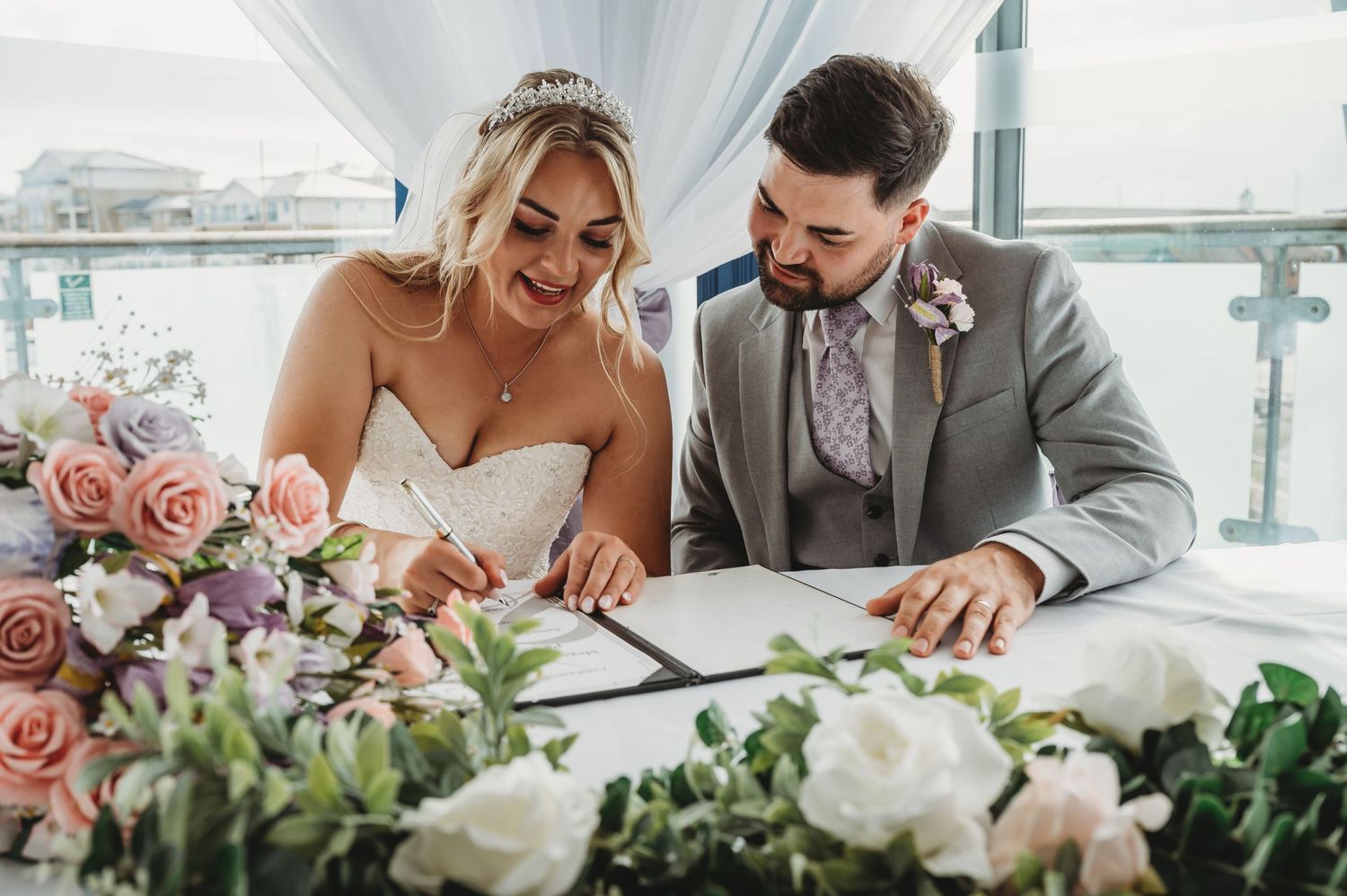 Happy couple sharing a laugh while signing wedding documents surrounded by pink and white flowers.