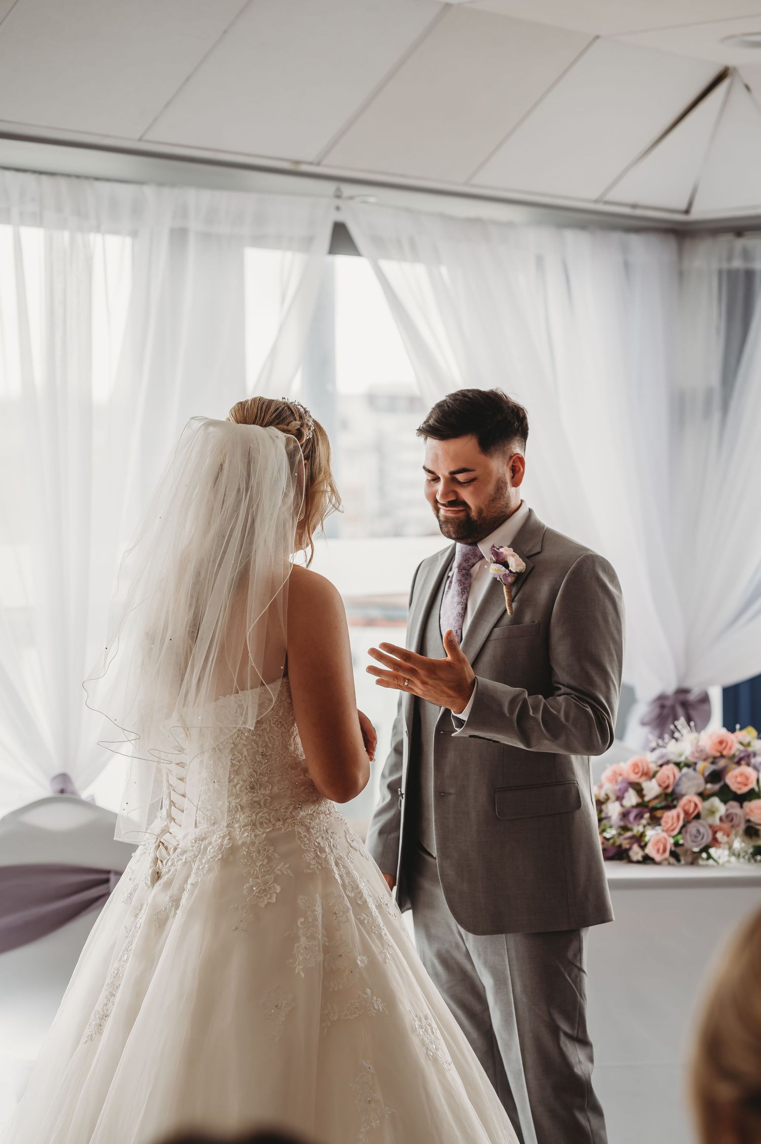 A couple exchanges vows during an intimate wedding ceremony under white sheer curtains with floral decor visible.