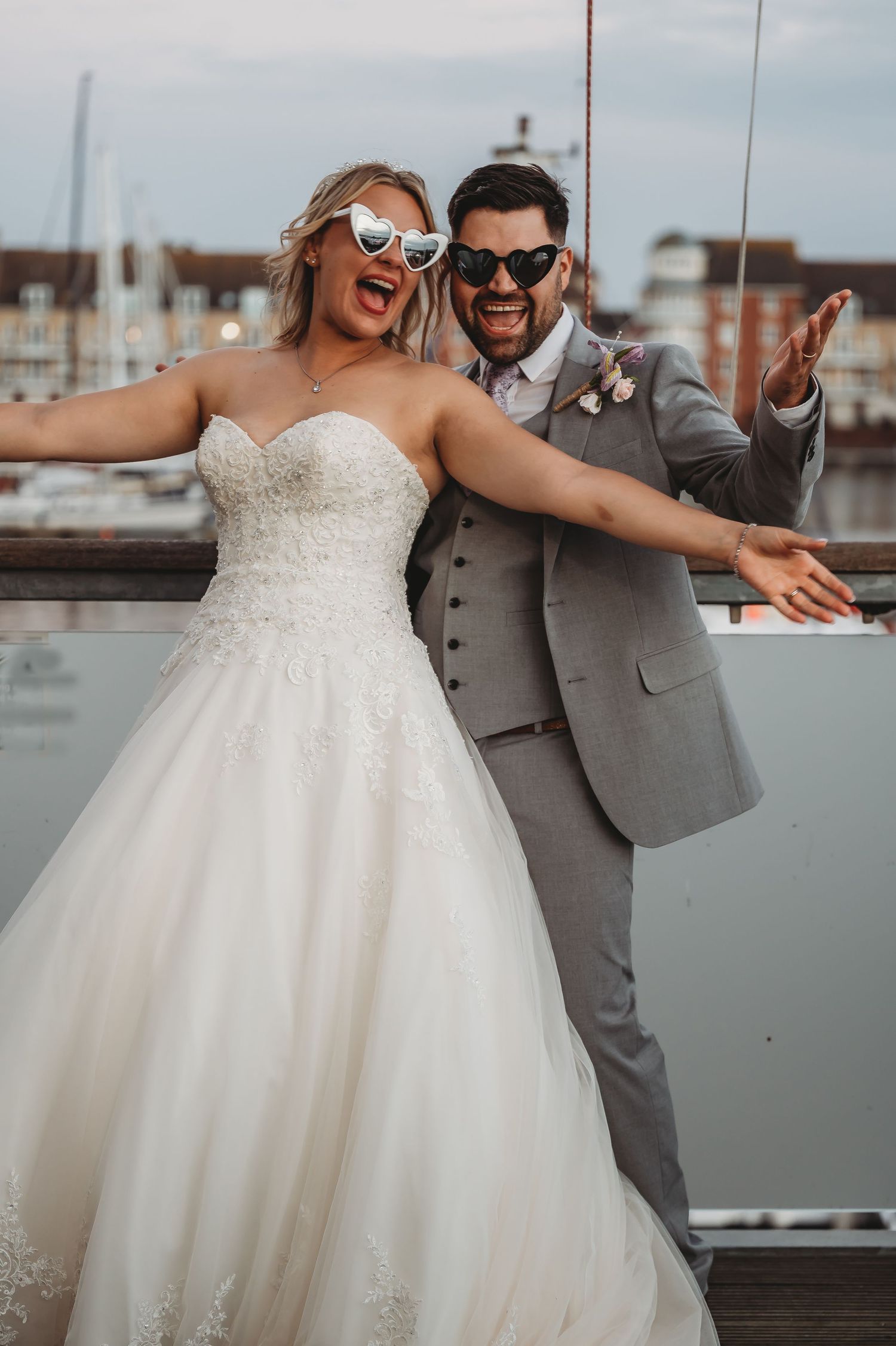 A joyful wedding couple celebrates on a dock wearing stylish sunglasses, with boats and buildings in the background.