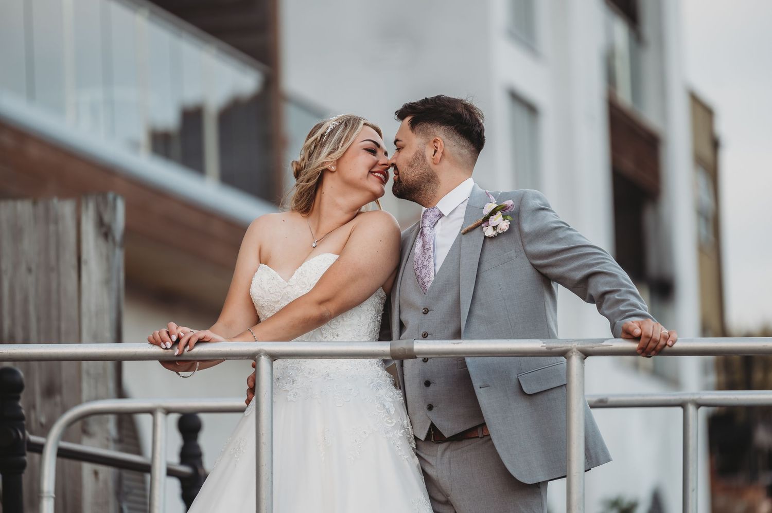 A couple in wedding attire share a romantic moment while leaning on a metal railing of a modern building.